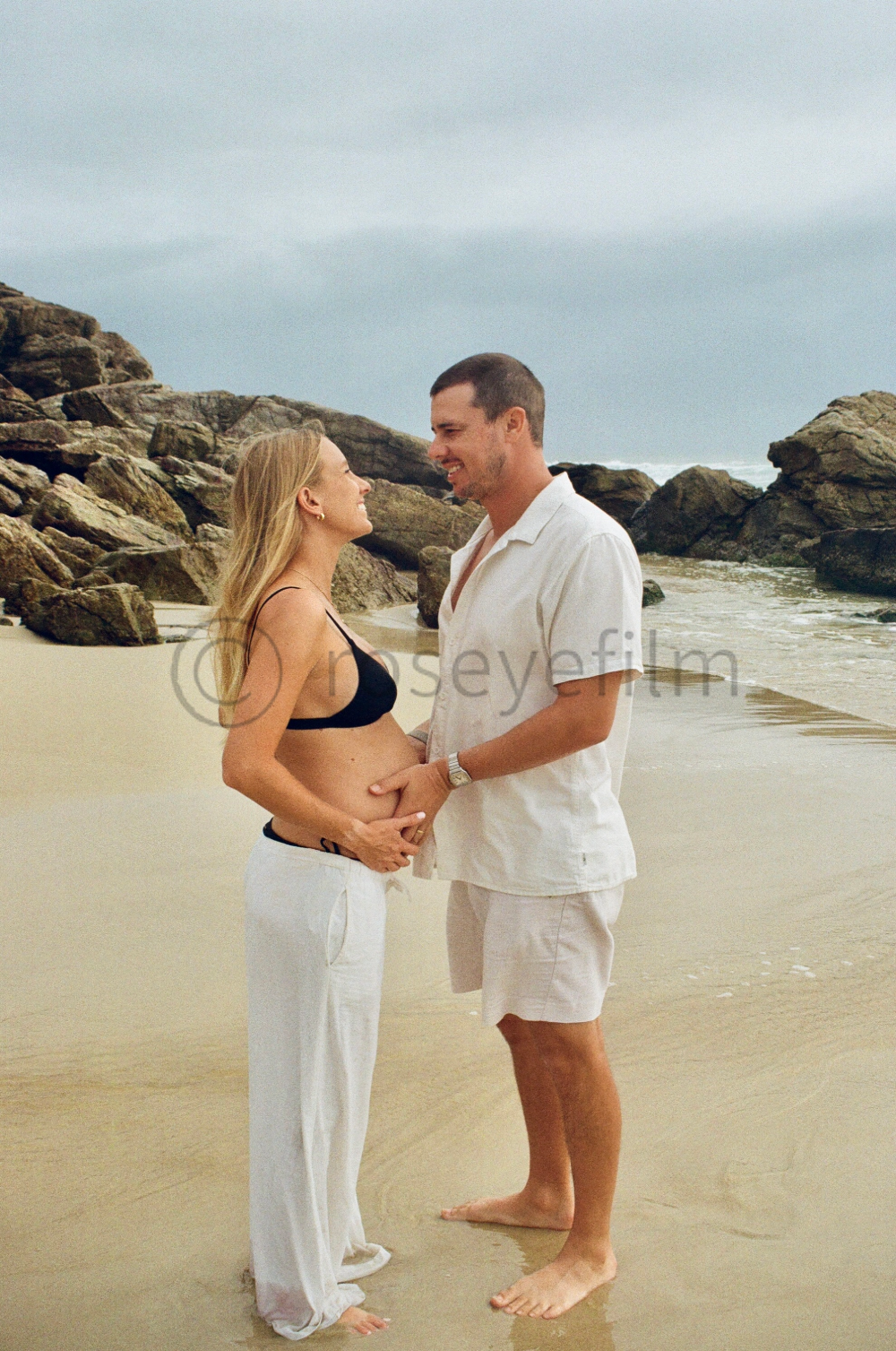 A pregnant woman and a man stand on the beach, holding hands and smiling at each other, with rocks and the ocean in the background.