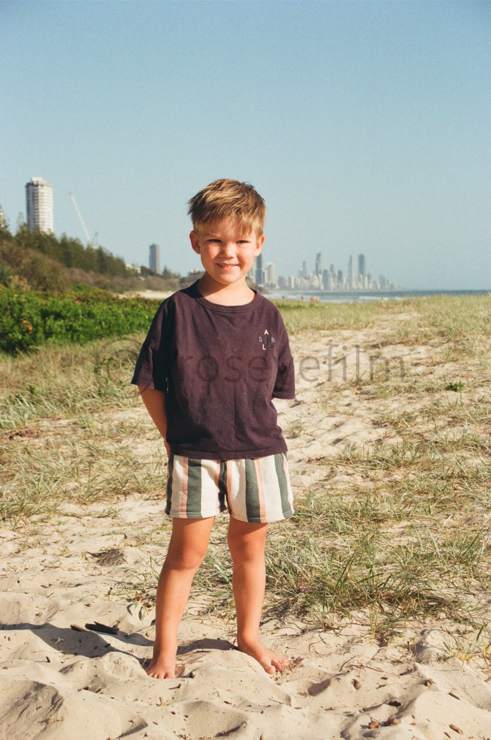 A young boy standing barefoot on a sandy beach with green grass, smiling at the camera, with city skyscrapers and a clear blue sky in the background.