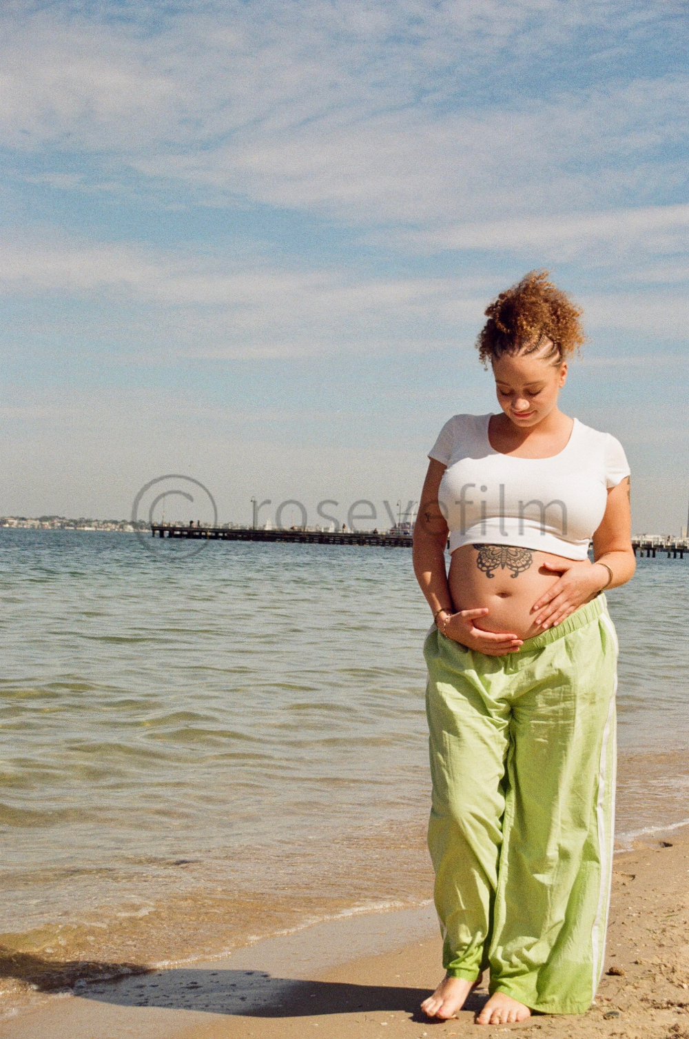 A pregnant woman standing on a sandy beach, holding her belly, with an ocean and a pier in the background, under a partly cloudy sky.