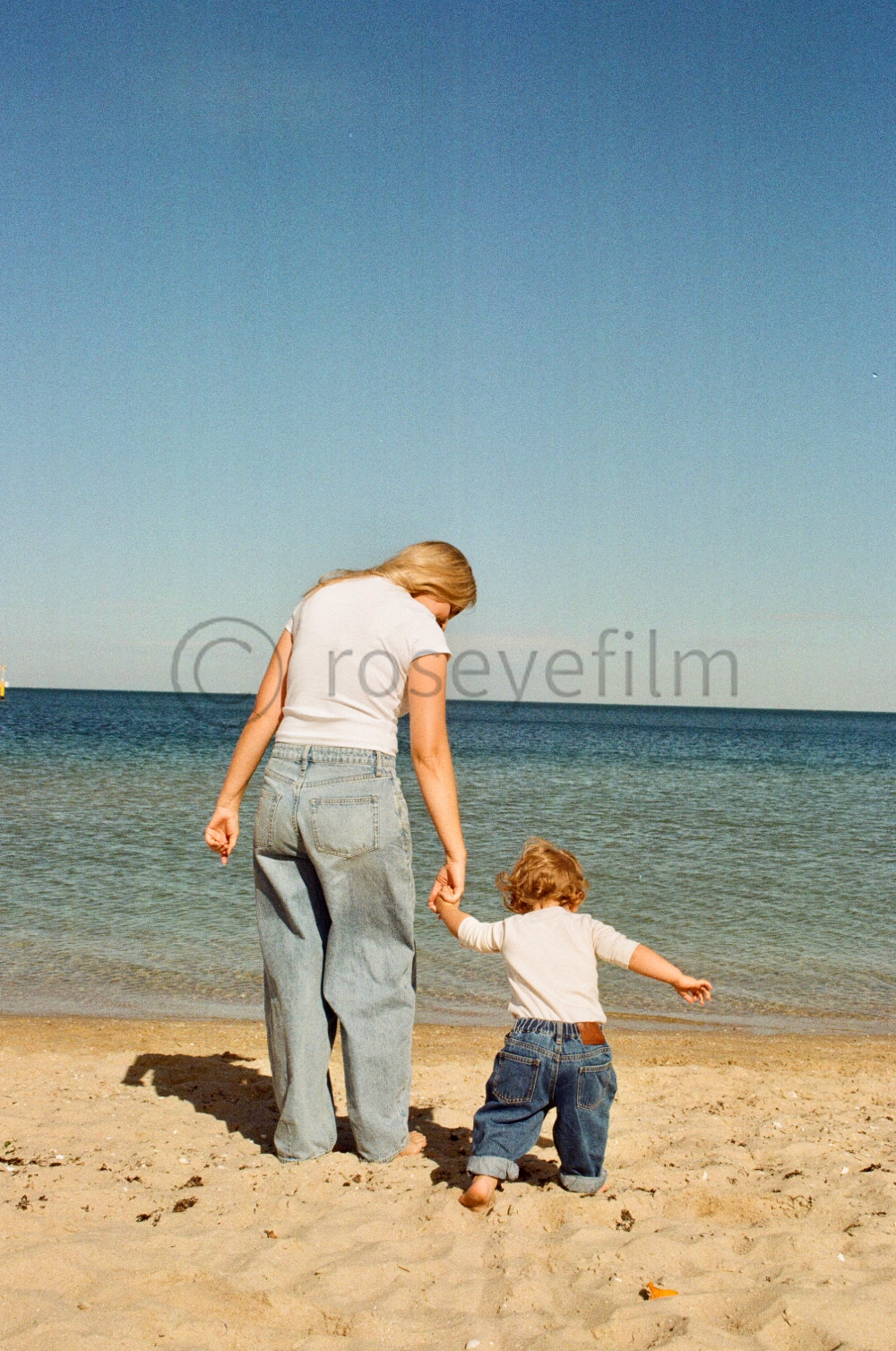 A woman and a young child walking hand in hand on a sandy beach by the ocean, with clear blue sky overhead.