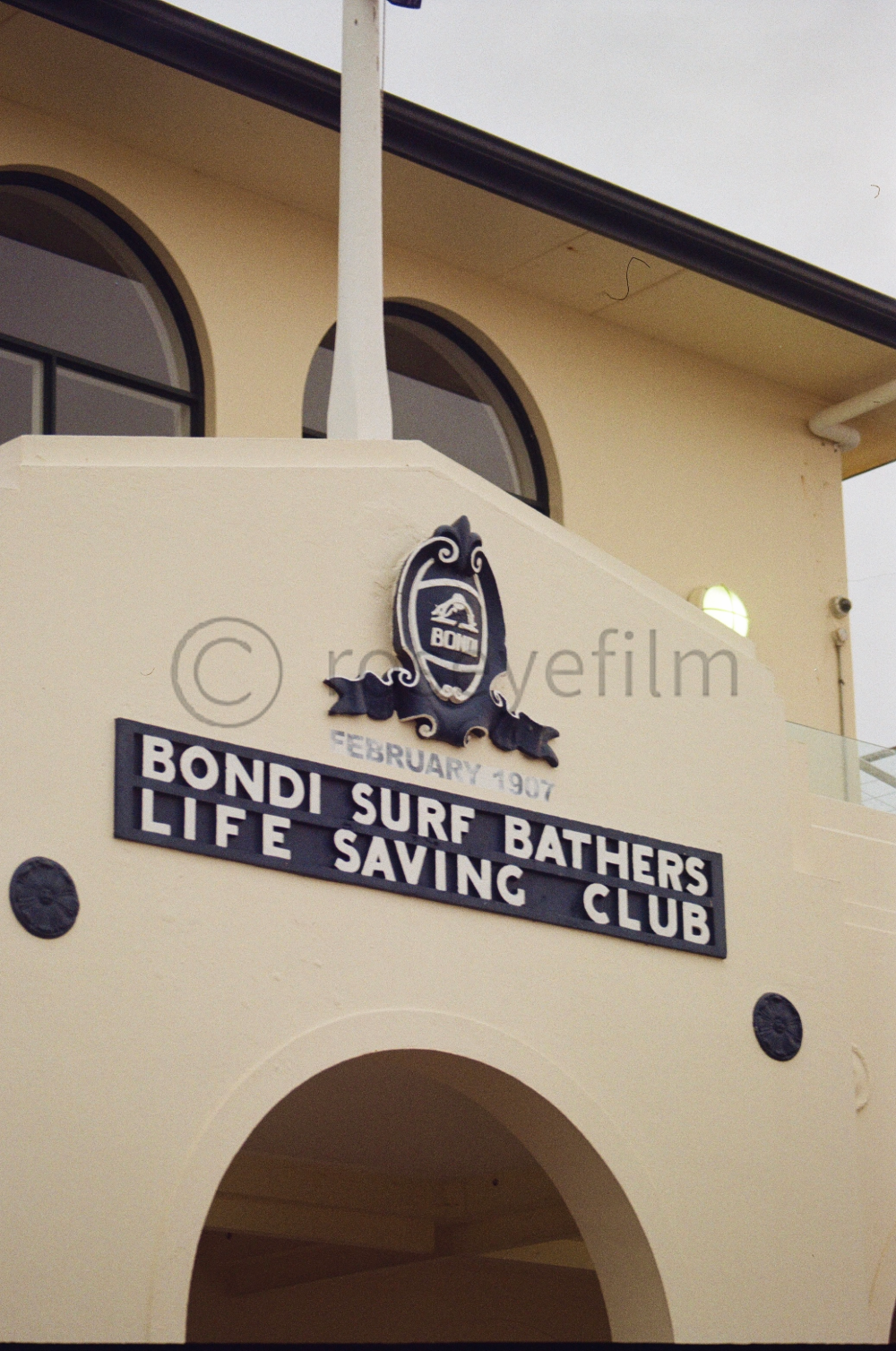 Sign for Bondi Surf Bathers Life Saving Club, established February 1907, on a building with arched windows.