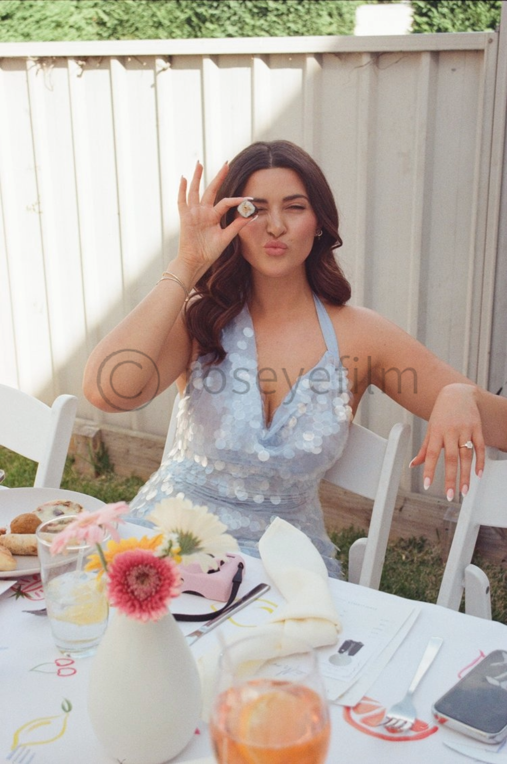 A woman with dark brown hair styled in loose waves, making a playful face while holding a cookie to her eye, sitting at an outdoor table with flowers, plates, and glasses, wearing a light blue, sequined dress with thin straps.