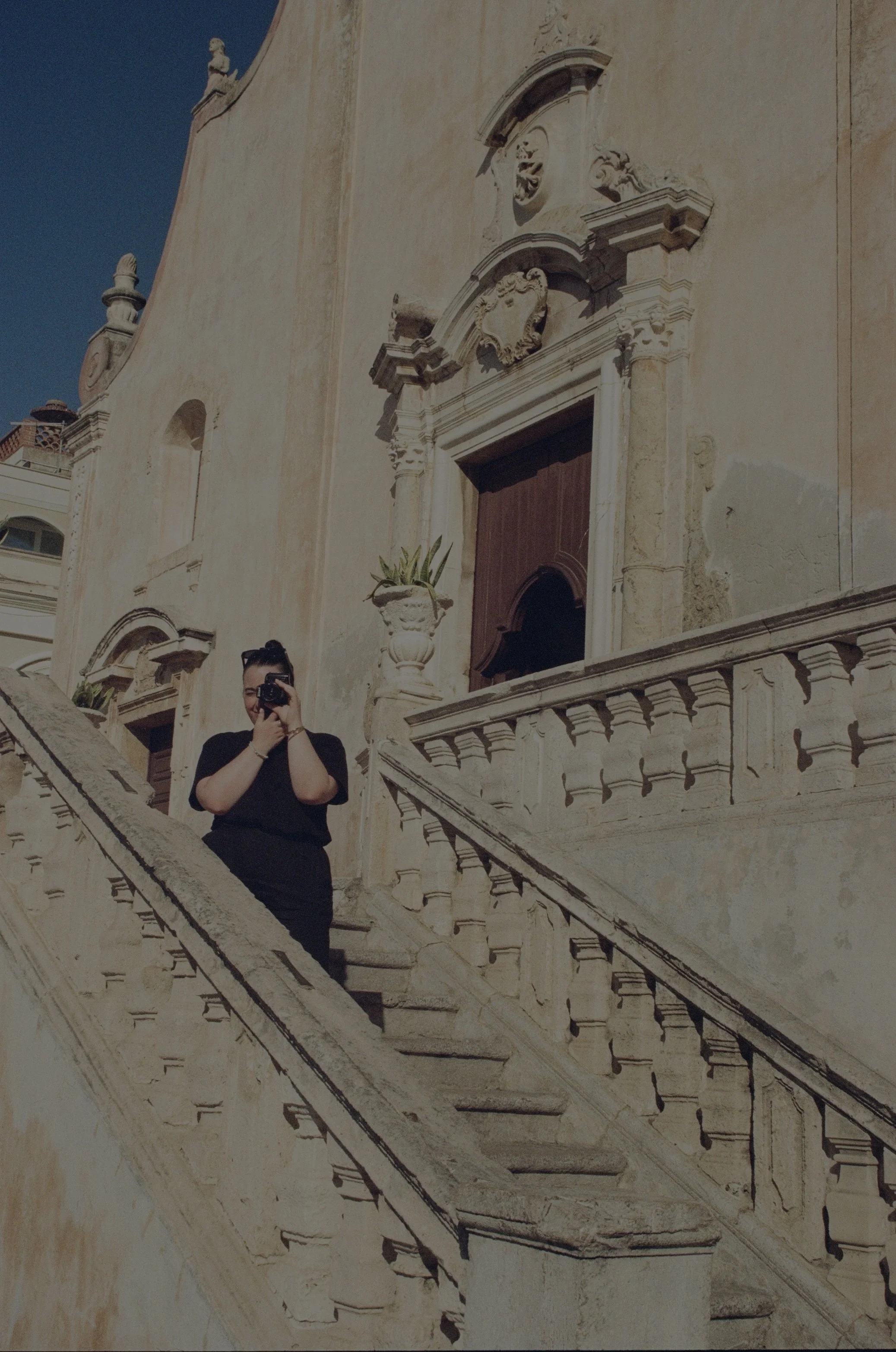 A woman standing on a staircase taking a photo with her camera, in front of an old building with ornate architectural details and sculptures.