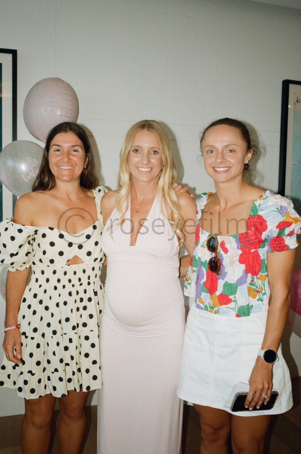 Three women standing at a celebration, smiling, with balloons in the background, one in a polka dot dress, one in a white dress, and one in a floral top and white skirt.