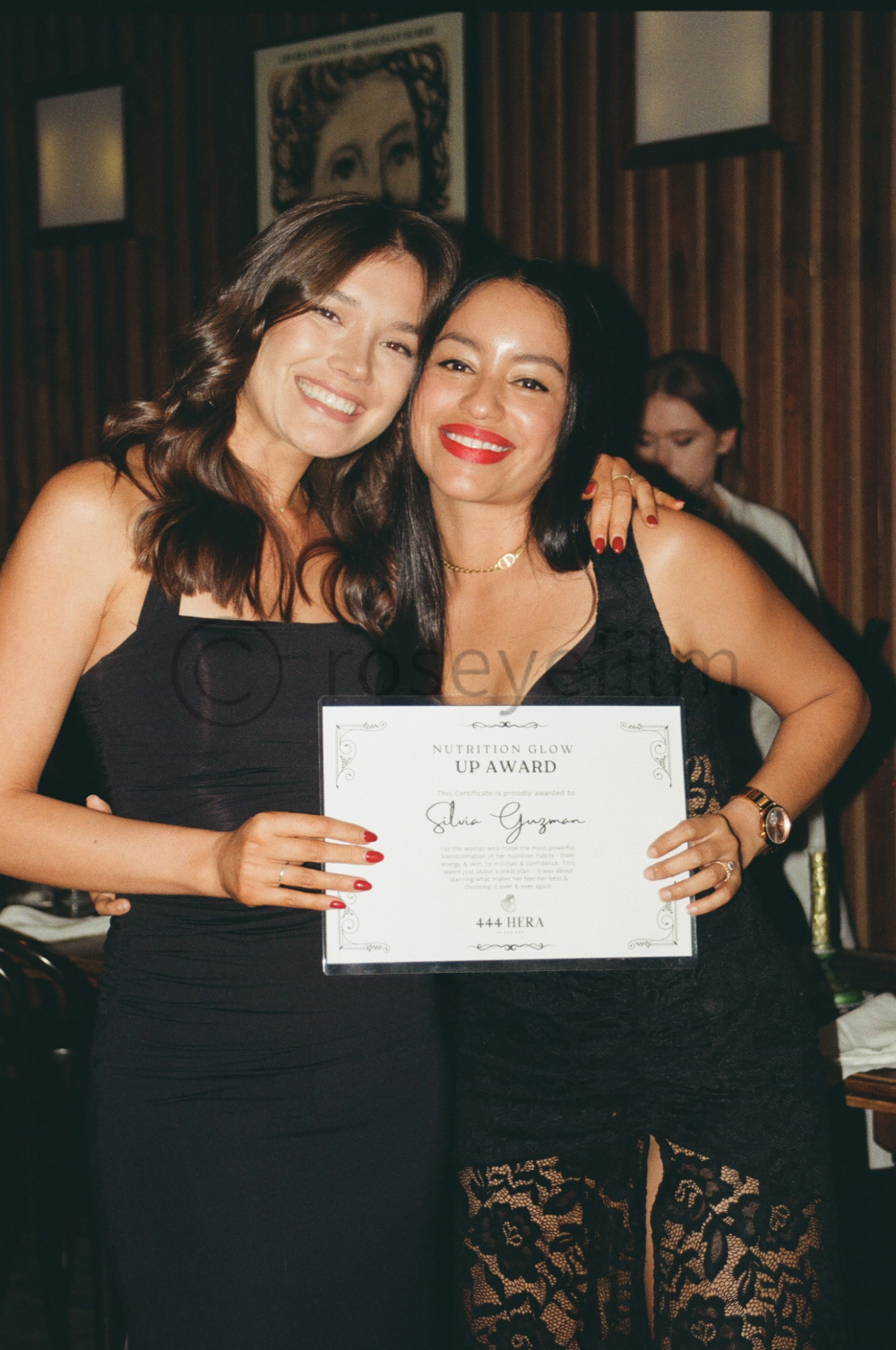 Two women smiling and holding a certificate at an awards event. They are dressed in black, with one wearing lace pants and the other in a black dress. The woman on the right has bright red lipstick, and both women have dark hair. The background featu