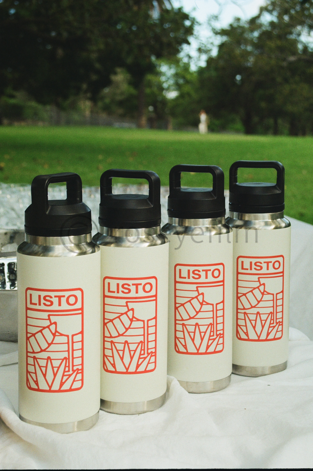 Four stainless steel water bottles with black caps and red labels are displayed outdoors on a white surface, with a blurred green park and trees in the background.
