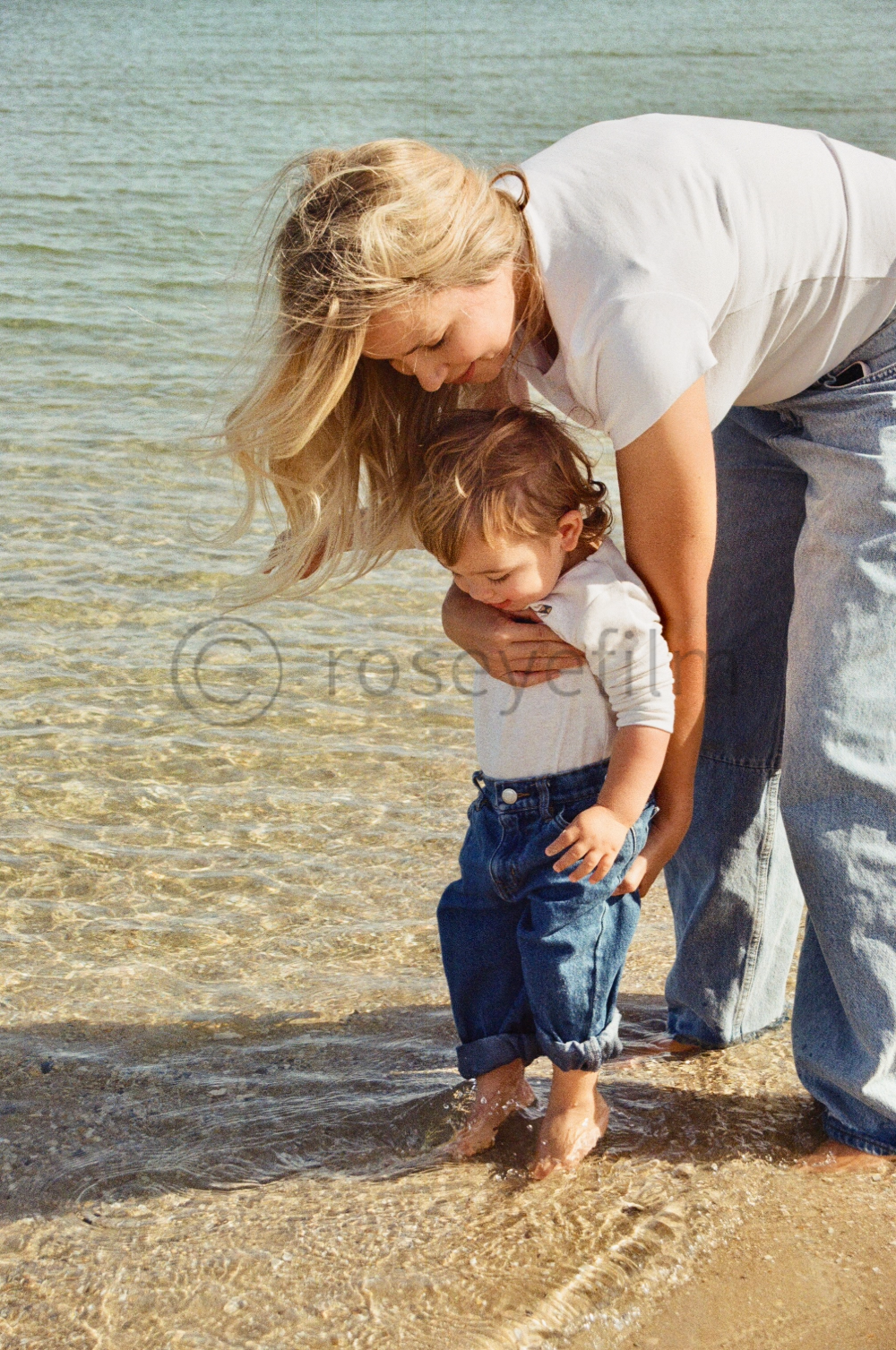 A woman helping a young boy stand in shallow water at the beach.