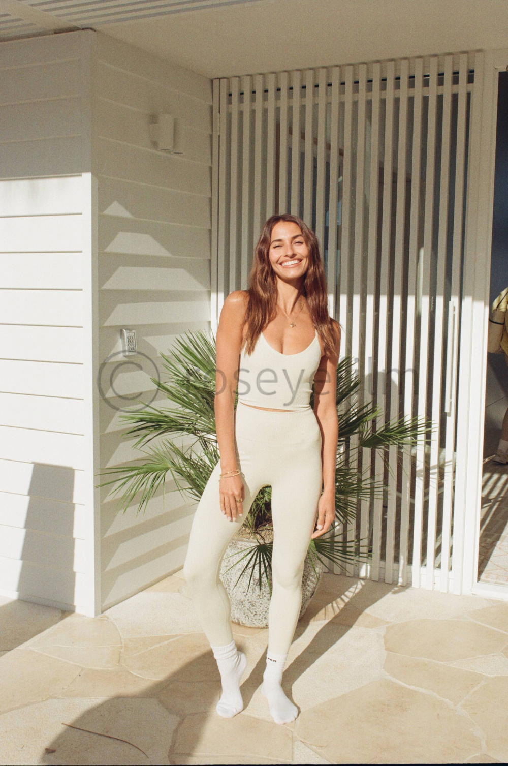A woman with long, wavy brown hair smiling outdoors, wearing a white sports bra and matching leggings, standing in front of a potted palm plant on a sunny patio.
