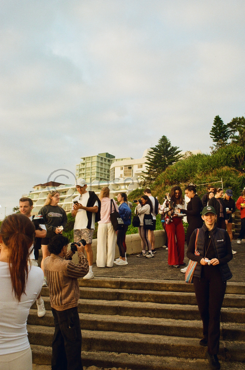 Group of people gathered outdoors on steps, some taking photos, chatting, and using smartphones, with buildings and trees in the background.