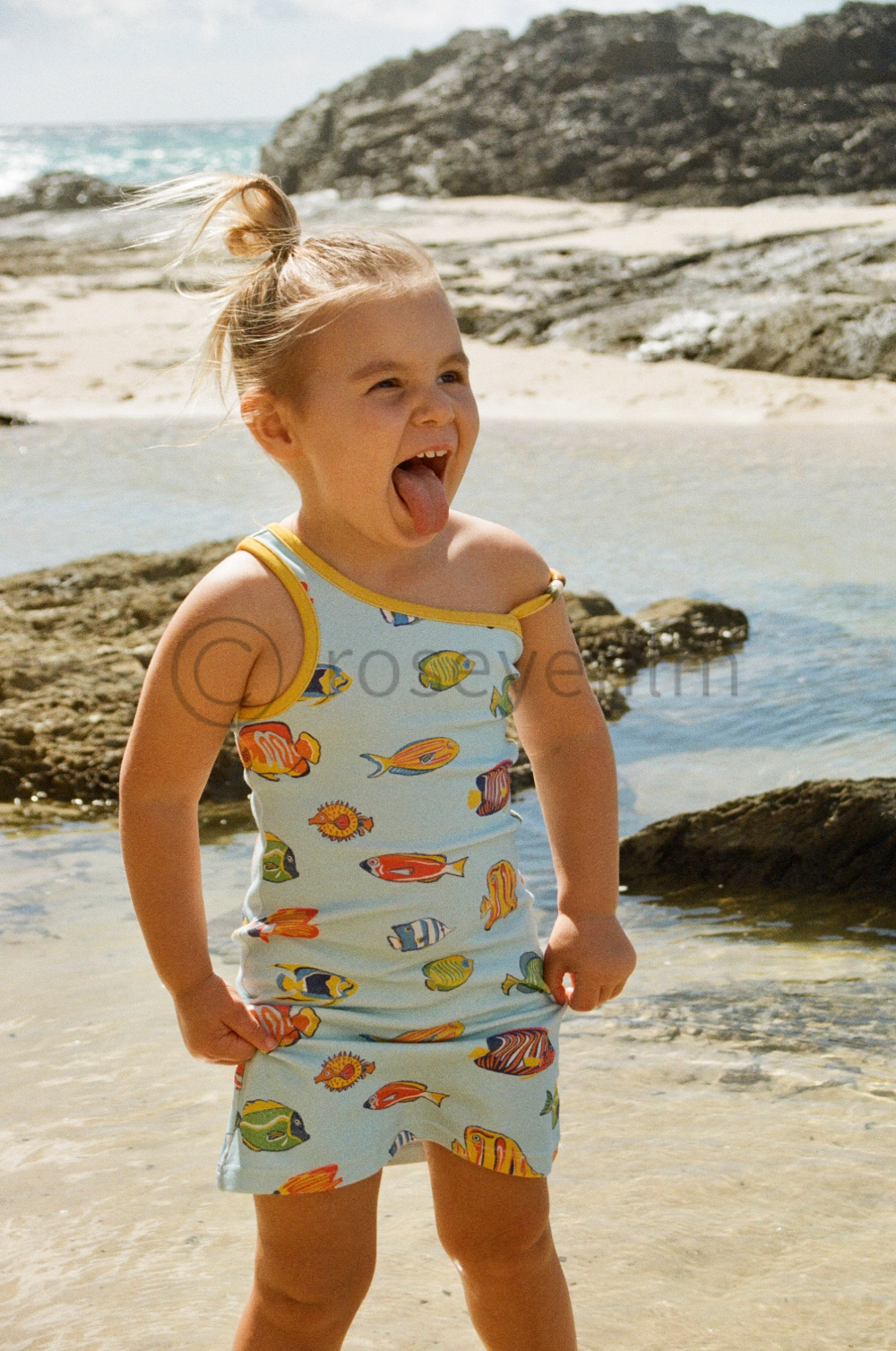 A young girl on the beach sticking out her tongue, wearing a sleeveless top and shorts with colorful fish patterns, near rocks and water.