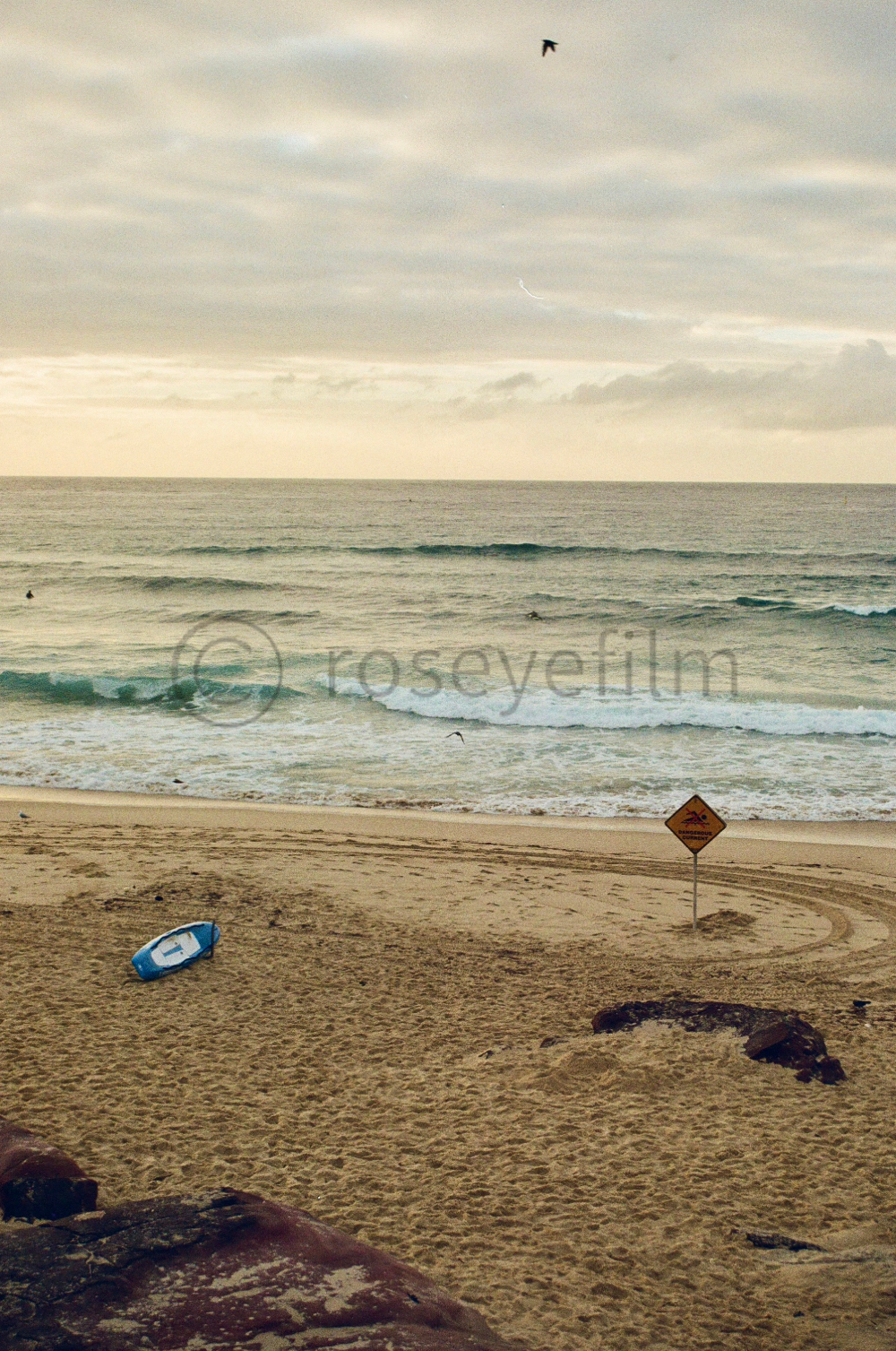 A beach scene with sand, waves, a few seagulls flying, a sign on the sand warning about strong currents, and a small blue boat lying on the beach.