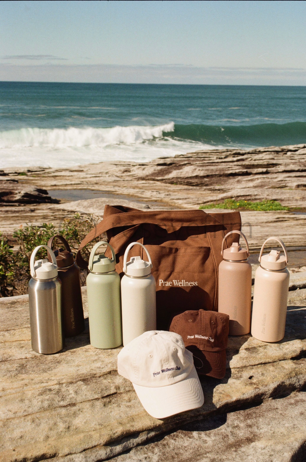 Assorted reusable bottles, a bag, caps, and a hat labeled 'Prae Wellness' and 'Prae Wellness Club' on a rocky surface near the beach with waves in the background.
