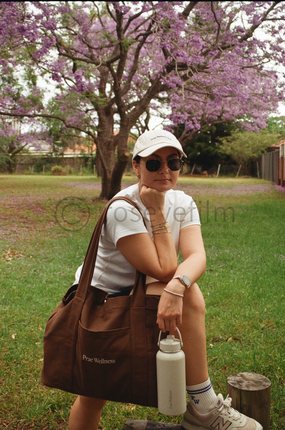 A woman sitting on a wooden post in a park with a large pink blooming tree in the background. She is wearing a white hat, sunglasses, a white shirt, beige shorts, and white sneakers. She has a large brown tote bag with the words "Prae Wellness" and i