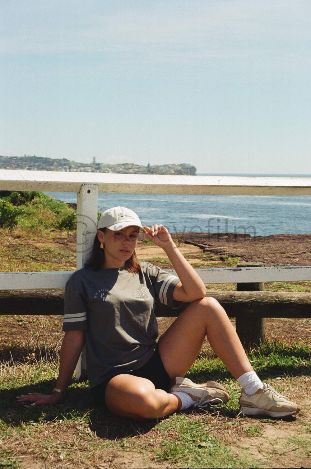A young woman sitting on the grass by a beach, wearing a gray t-shirt, white cap, white sneakers, and white socks. She is seated with one knee up and her hand adjusting her cap, with water, rocky shoreline, and land visible in the background.