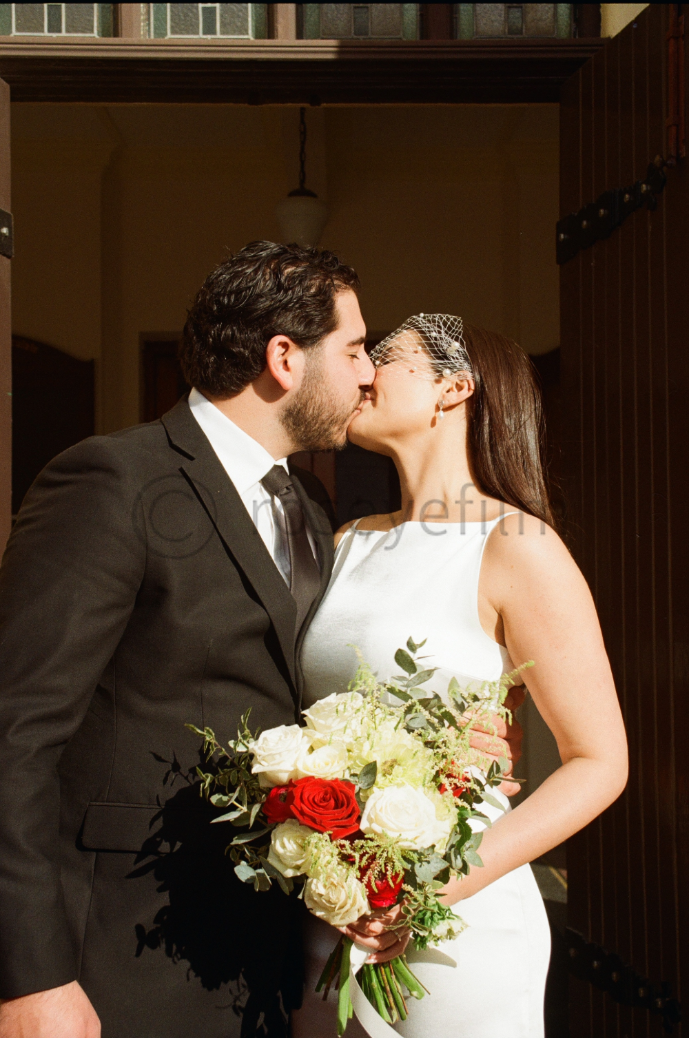 A bride and groom sharing a kiss during their wedding. The groom is in a black suit and tie, and the bride is wearing a white wedding dress with an elegant netted veil. She is holding a bouquet of white, red, and green flowers.