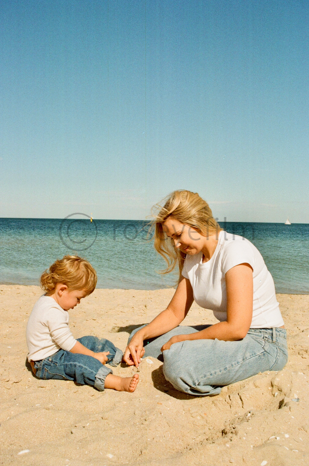 A woman and a young girl sitting on the sandy beach, playing with shells near the water with sailboats visible in the distance under a clear blue sky.