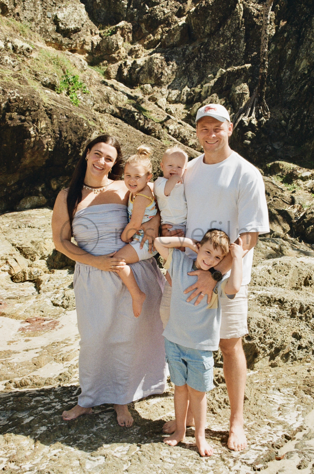 Family of six posing together on a rocky beach with large rocks and cliffs in the background, smiling at the camera.