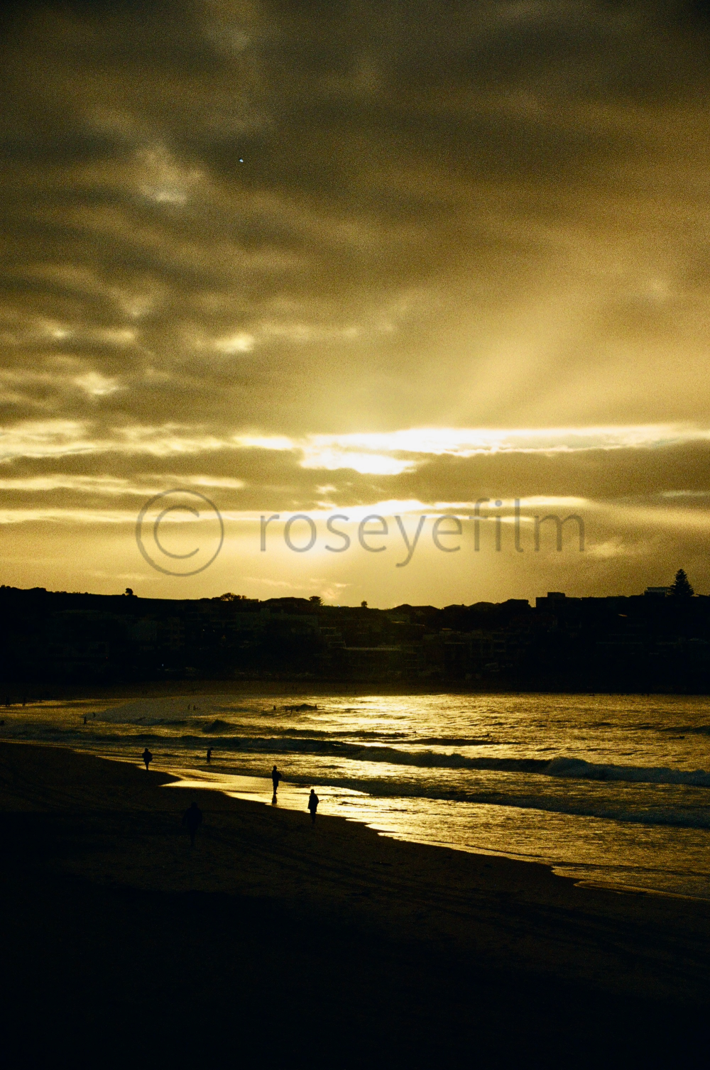 A cloudy sunset over a beach with silhouettes of people walking along the shore and houses in the distance.