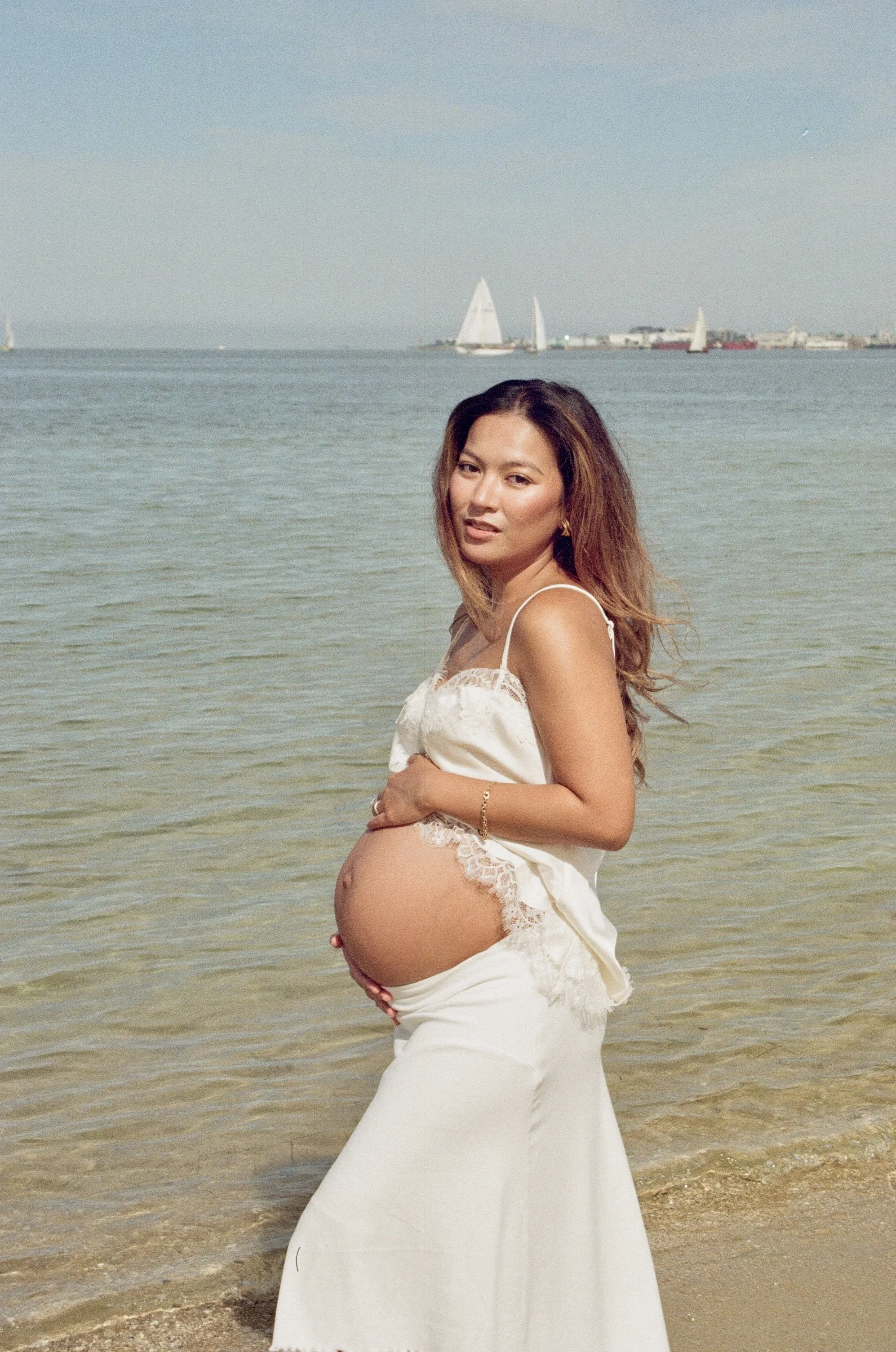 A pregnant woman in a white dress standing on the beach with water and sailboats in the background.