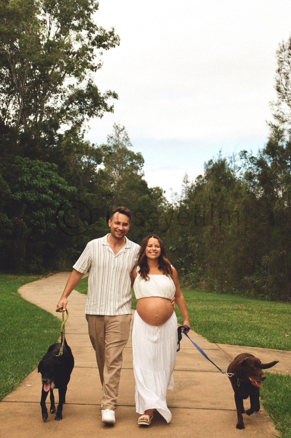 A pregnant woman and a man are walking on a park trail, each holding a dog on a leash, with trees and grass around them on a cloudy day.