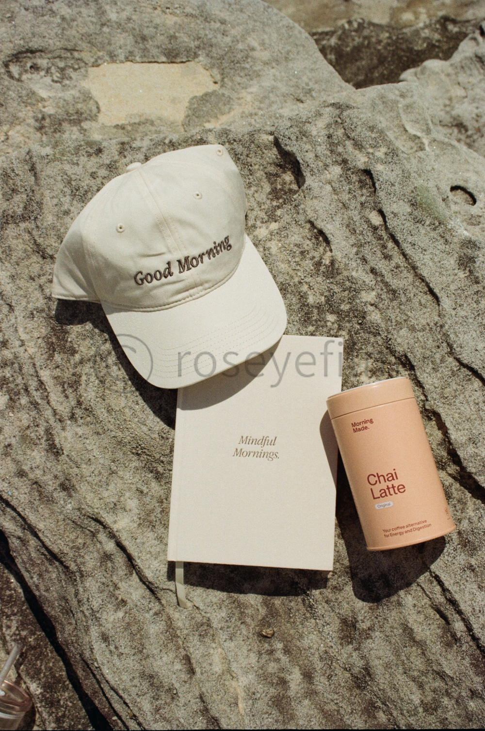A white baseball cap embroidered with 'Good Morning,' a pink container labeled 'Chai Latte,' and an open minimalist booklet on a rocky surface.
