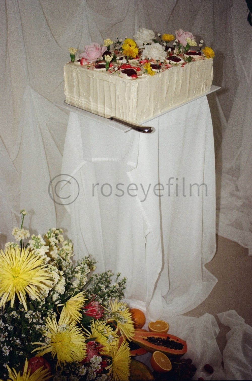 A rectangular white cake decorated with flowers on a stand, with a bouquet of yellow flowers, papaya, orange, and cherries at the base.