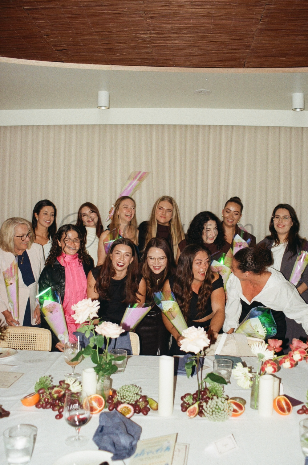 Group of women celebrating at a dinner table, holding bouquets, smiling, with floral centerpieces and food on the table.