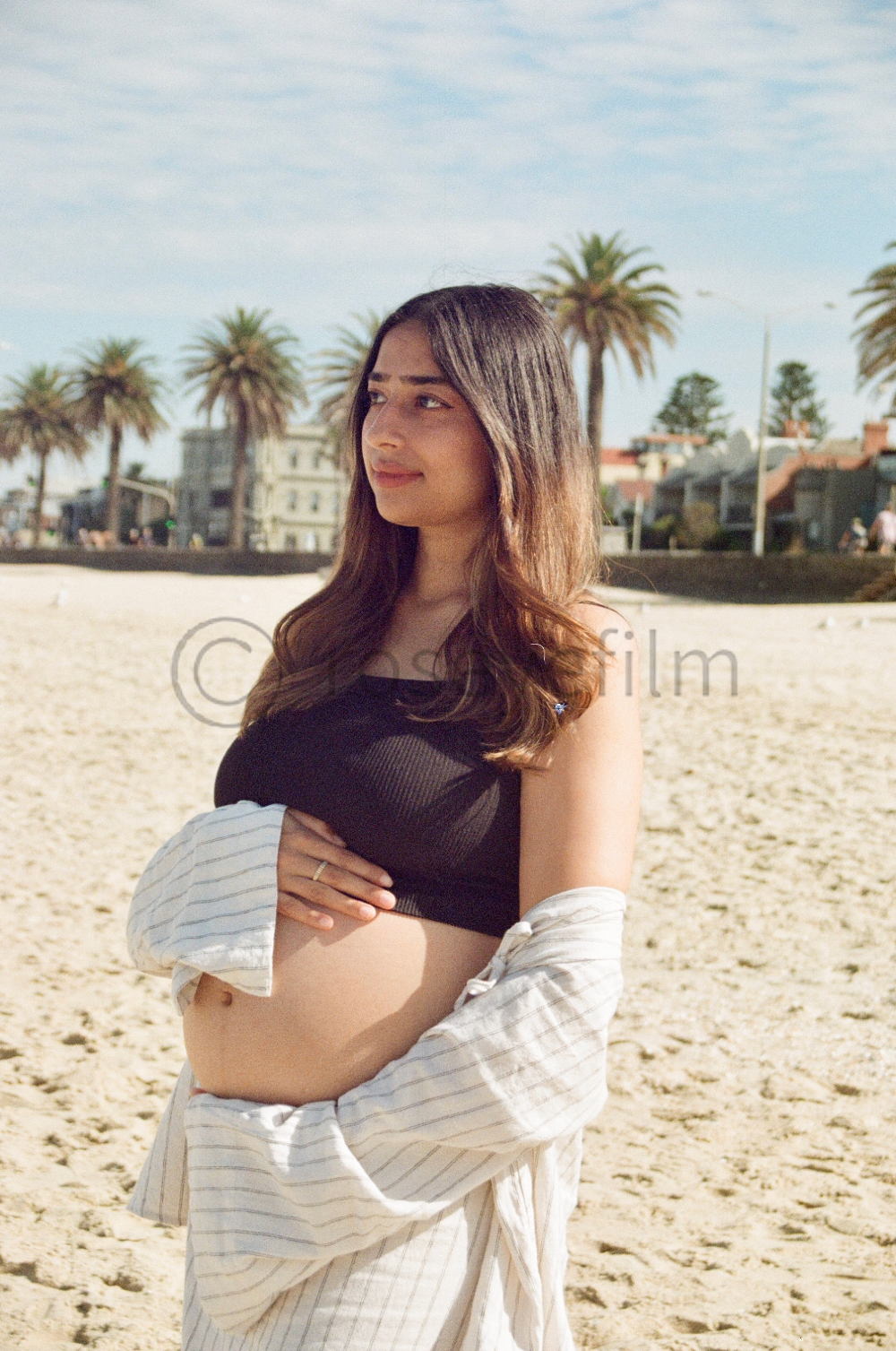 A pregnant woman stands on a sandy beach, with palm trees and buildings in the background, looking contemplative.