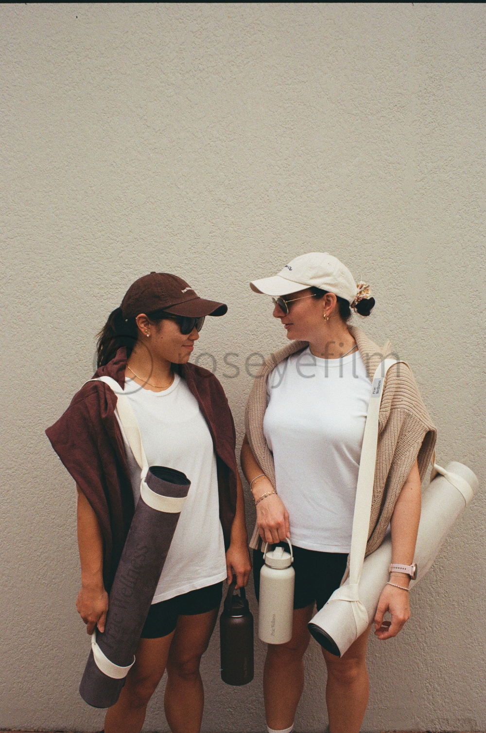 Two women dressed in athletic wear, wearing caps and sunglasses, standing against a plain wall, each holding a water bottle and carrying yoga mats.