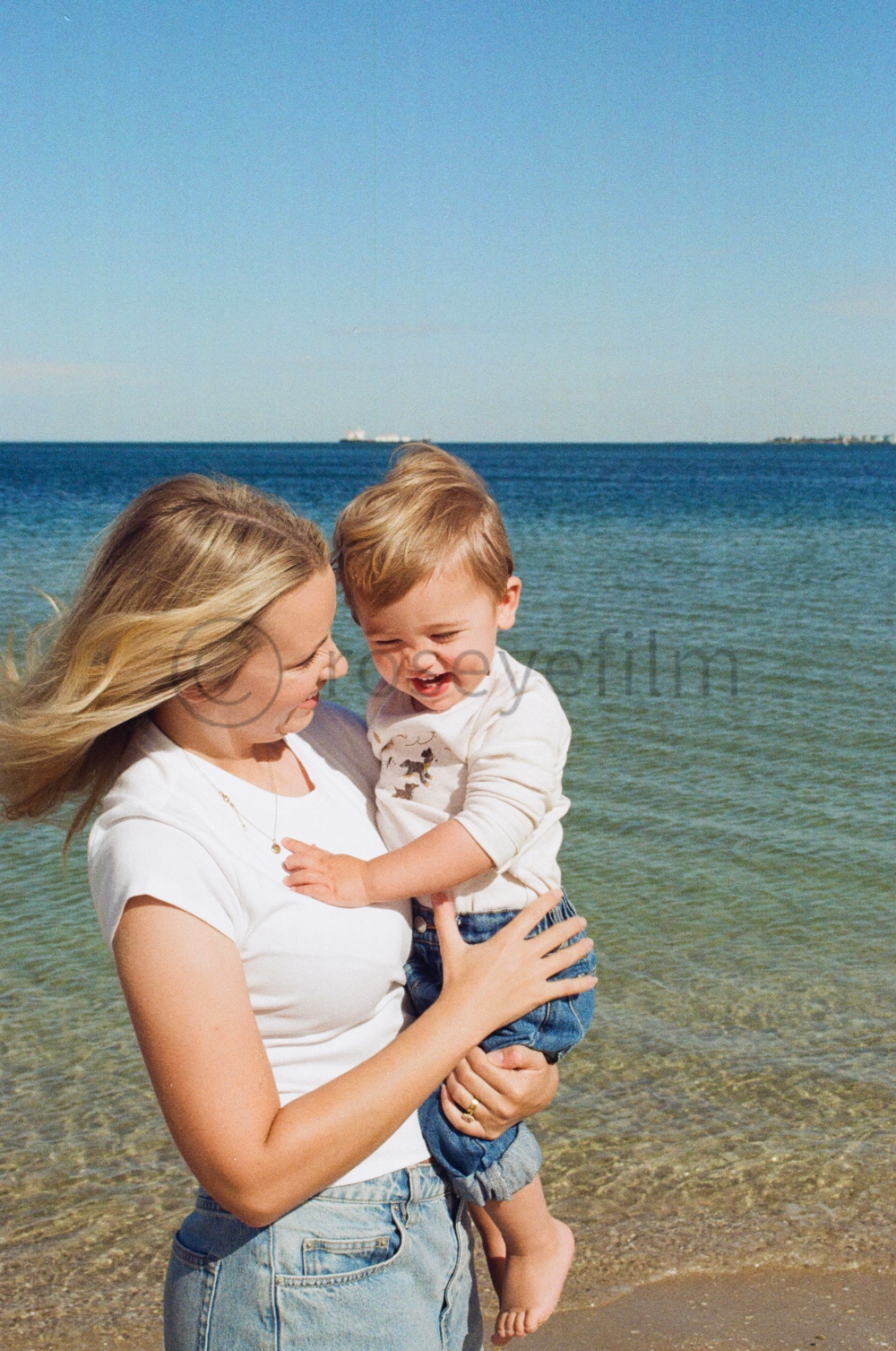 A woman holding a laughing young boy on a beach during daytime, with the ocean and blue sky in the background.