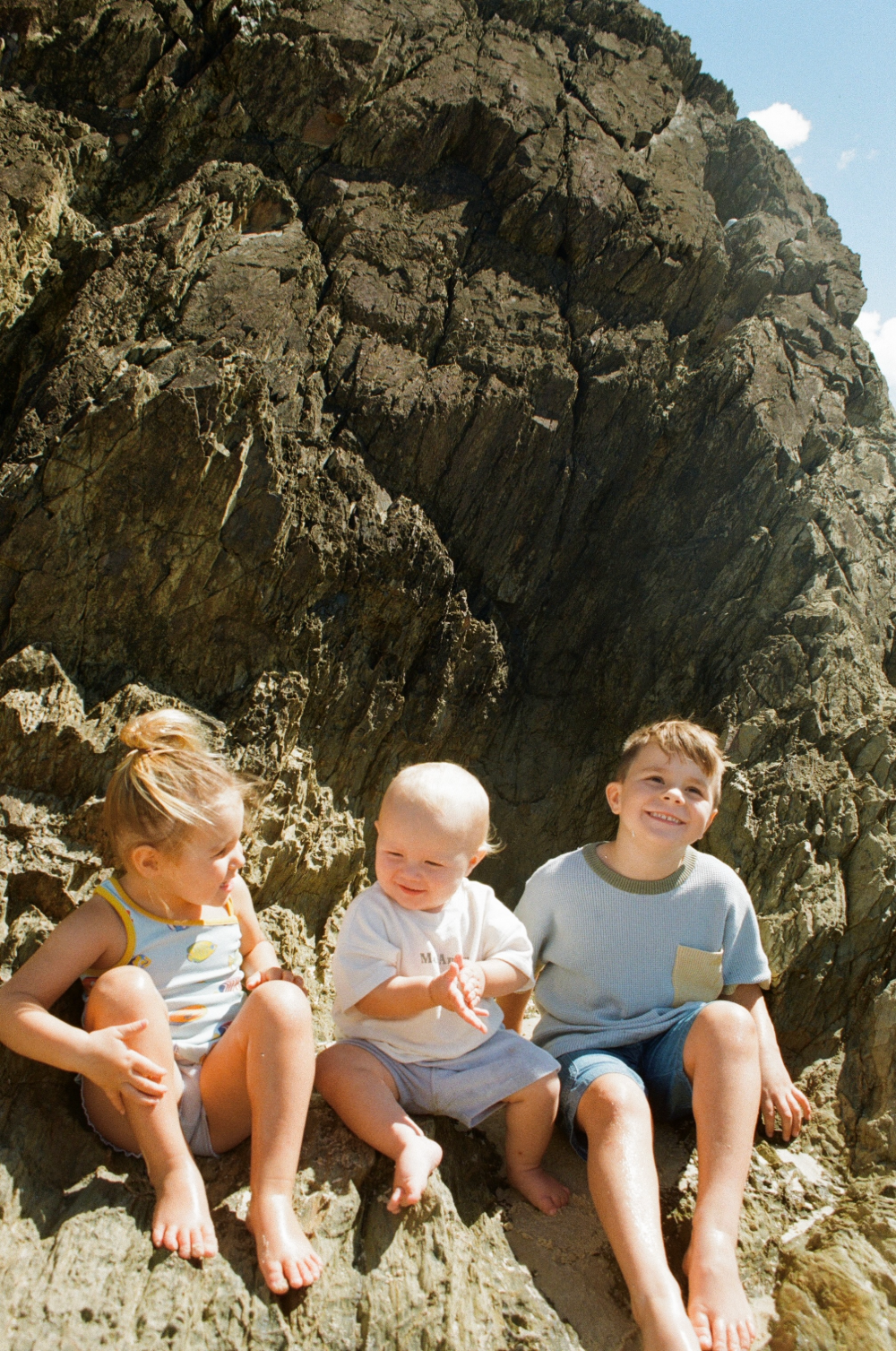 Three children sitting on rocks near a large rocky cliff with a small patch of blue sky and clouds visible above.