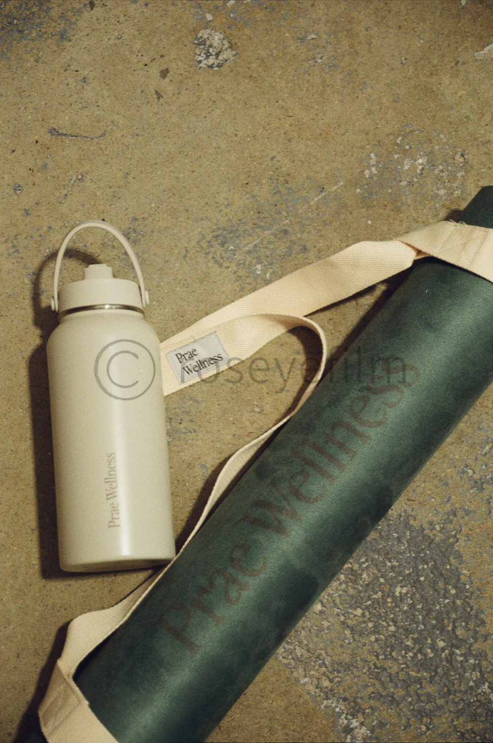A white water bottle and a black yoga mat with a beige strap lying on a concrete floor.