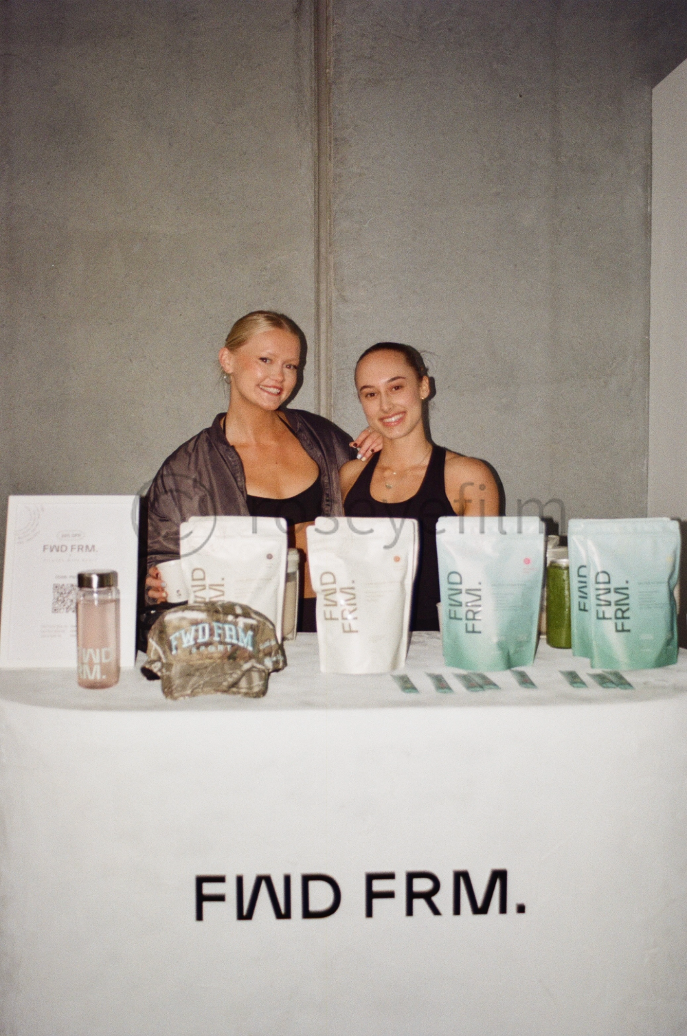 Two women standing behind a table with FWD FIRM branding, displaying various packaged products and promotional items.