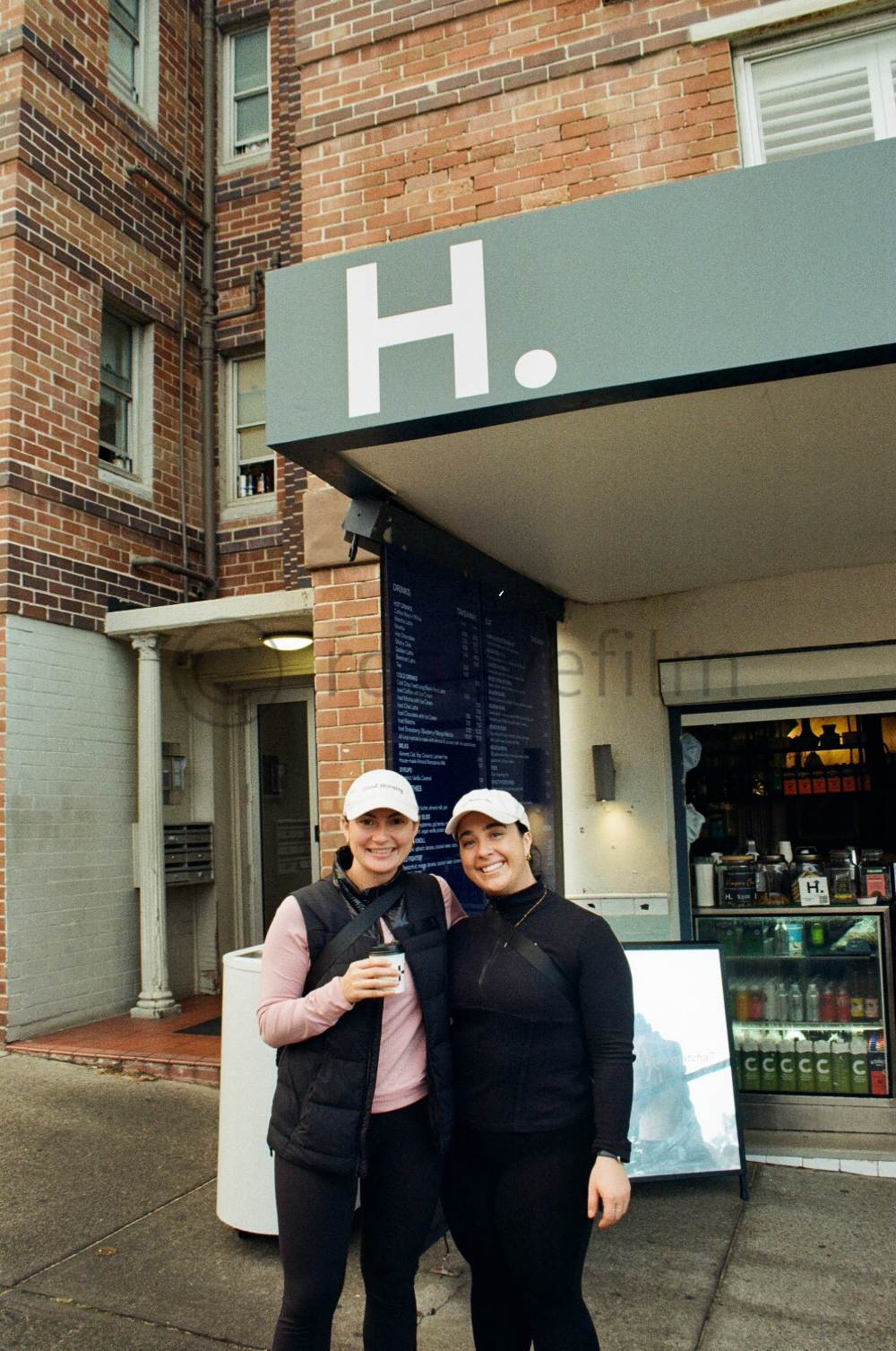 Two women smiling and standing outside a café or coffee shop, one holding a drink, in front of a brick building and a menu board with a prominent H. sign.