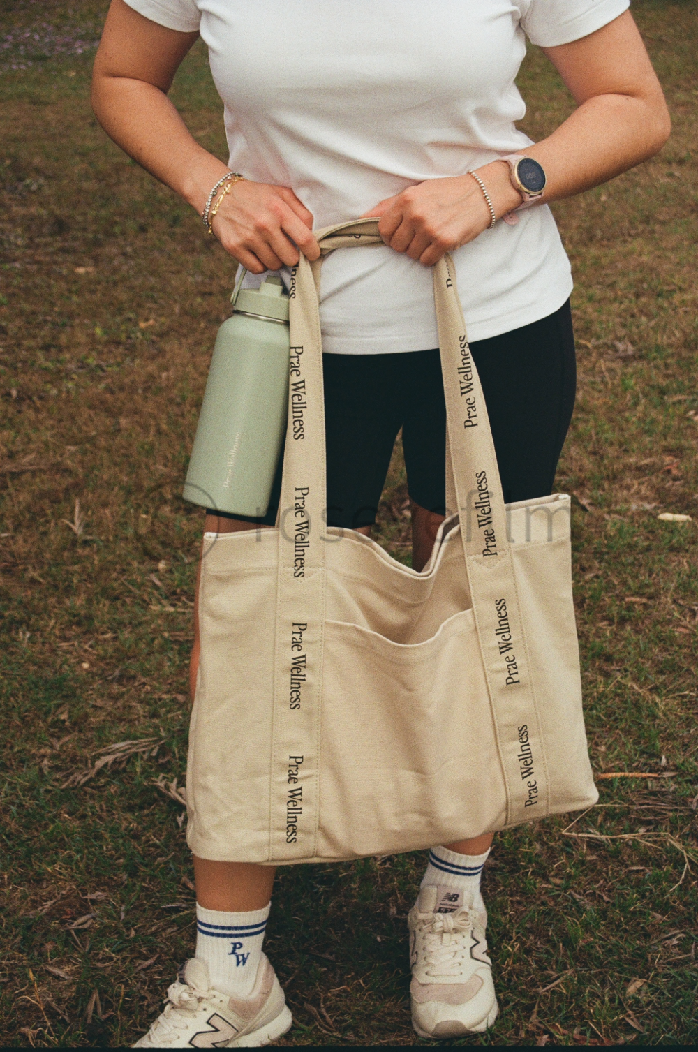 Person holding a beige tote bag with 'Prae Wellness' printed on the straps, in an outdoor setting with grass. The person is wearing white sneakers, white socks with blue stripes, black leggings, a white t-shirt, a smartwatch, and bracelets. They have
