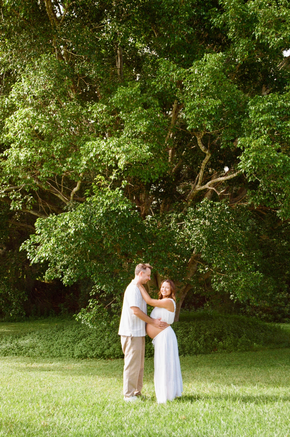 A pregnant woman and a man standing on grass under a large leafy tree, smiling at each other, with the woman touching her belly.