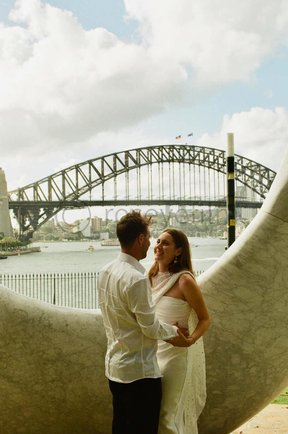 A couple standing close in front of a large artistic sculpture with the Sydney Harbour Bridge in the background, over the water with boats and city skyline.