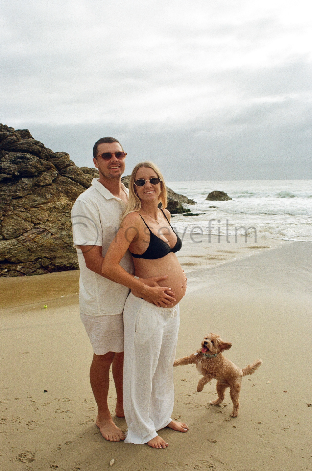A pregnant woman and her partner standing on a beach with a dog, ocean waves, and rocks in the background.