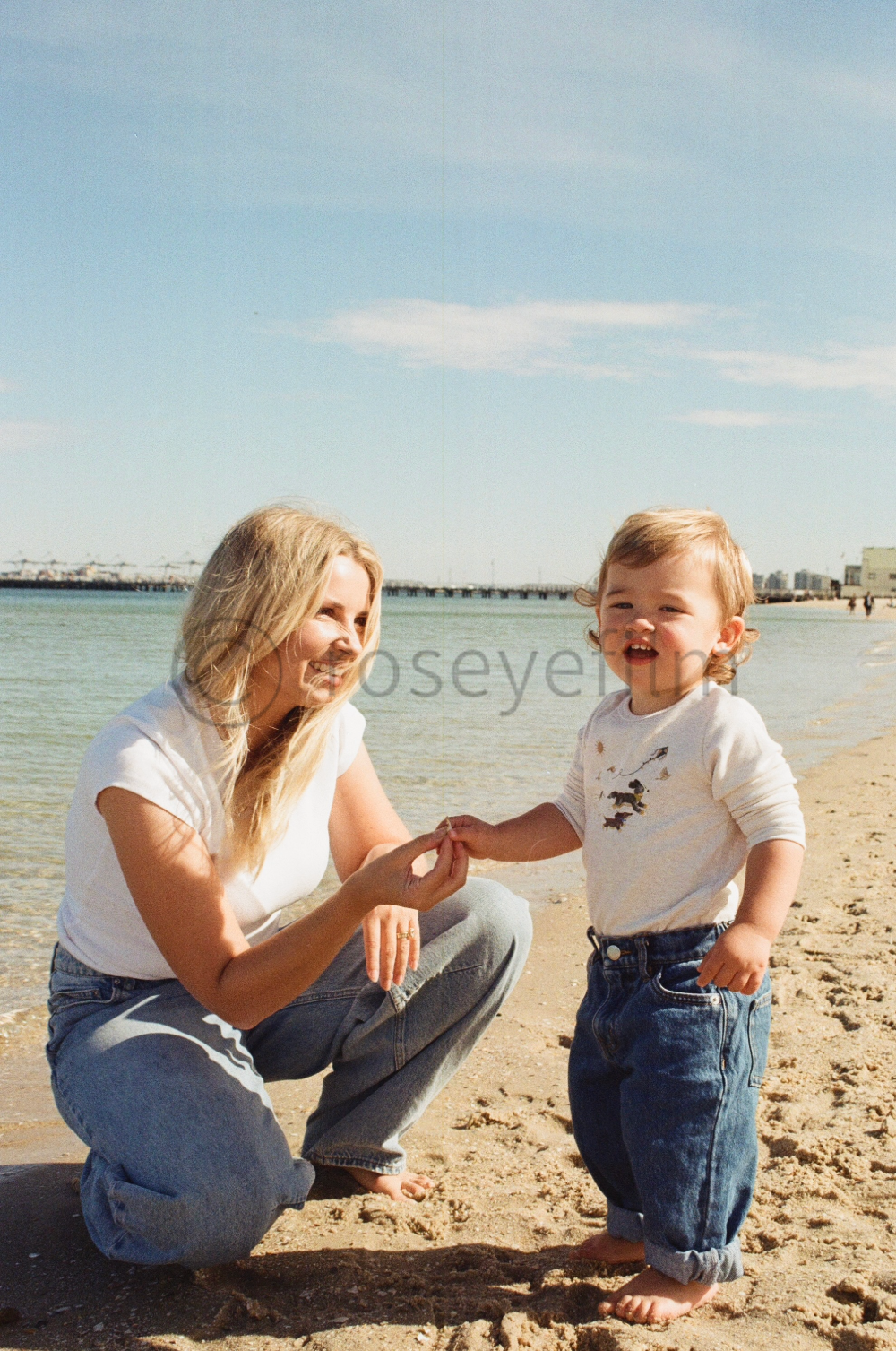 A woman and a young child with blond hair at the beach, holding hands, with the ocean and a pier in the background.
