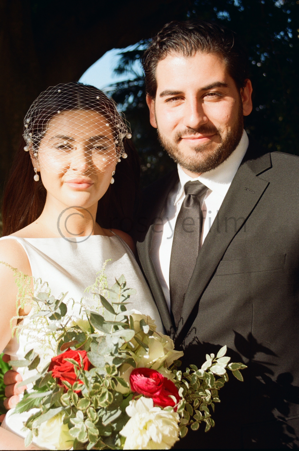 A bride holding a bouquet of red, white, and green flowers, standing next to a groom dressed in a black suit and tie, both smiling outdoors.