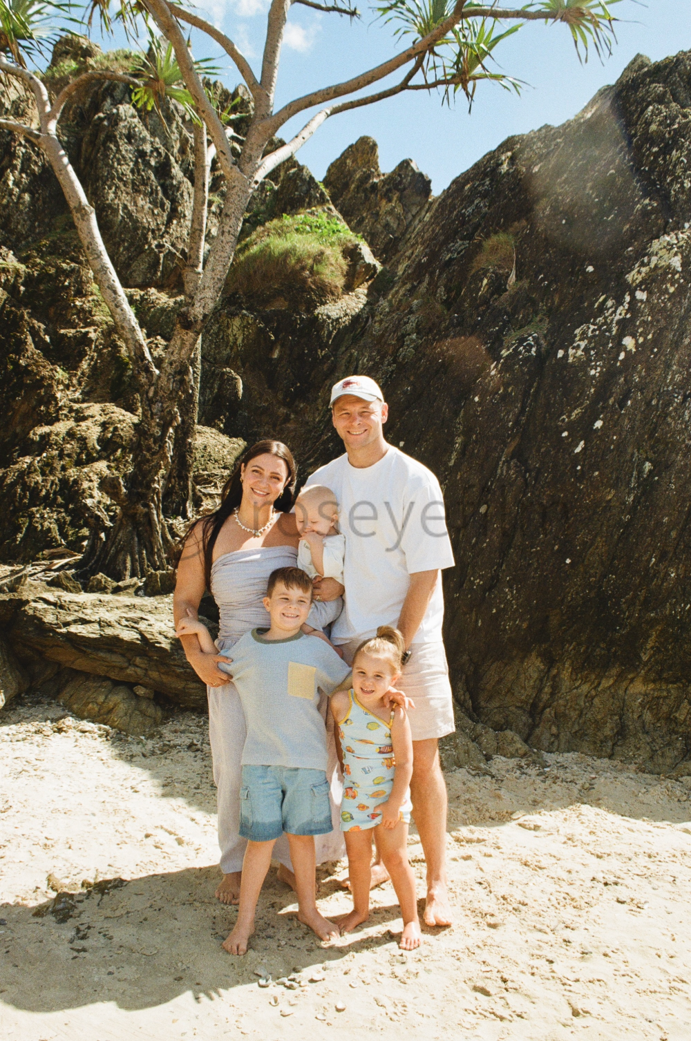 A family of six posing on a sandy beach with large rock formations and a small tree in the background, under a sunny sky.