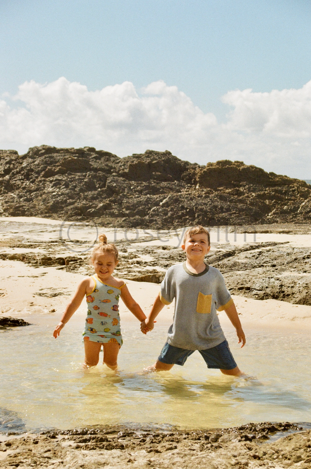 Two children, a girl and a boy, holding hands and smiling while standing in shallow water at the beach with rocks and a cloudy sky in the background.