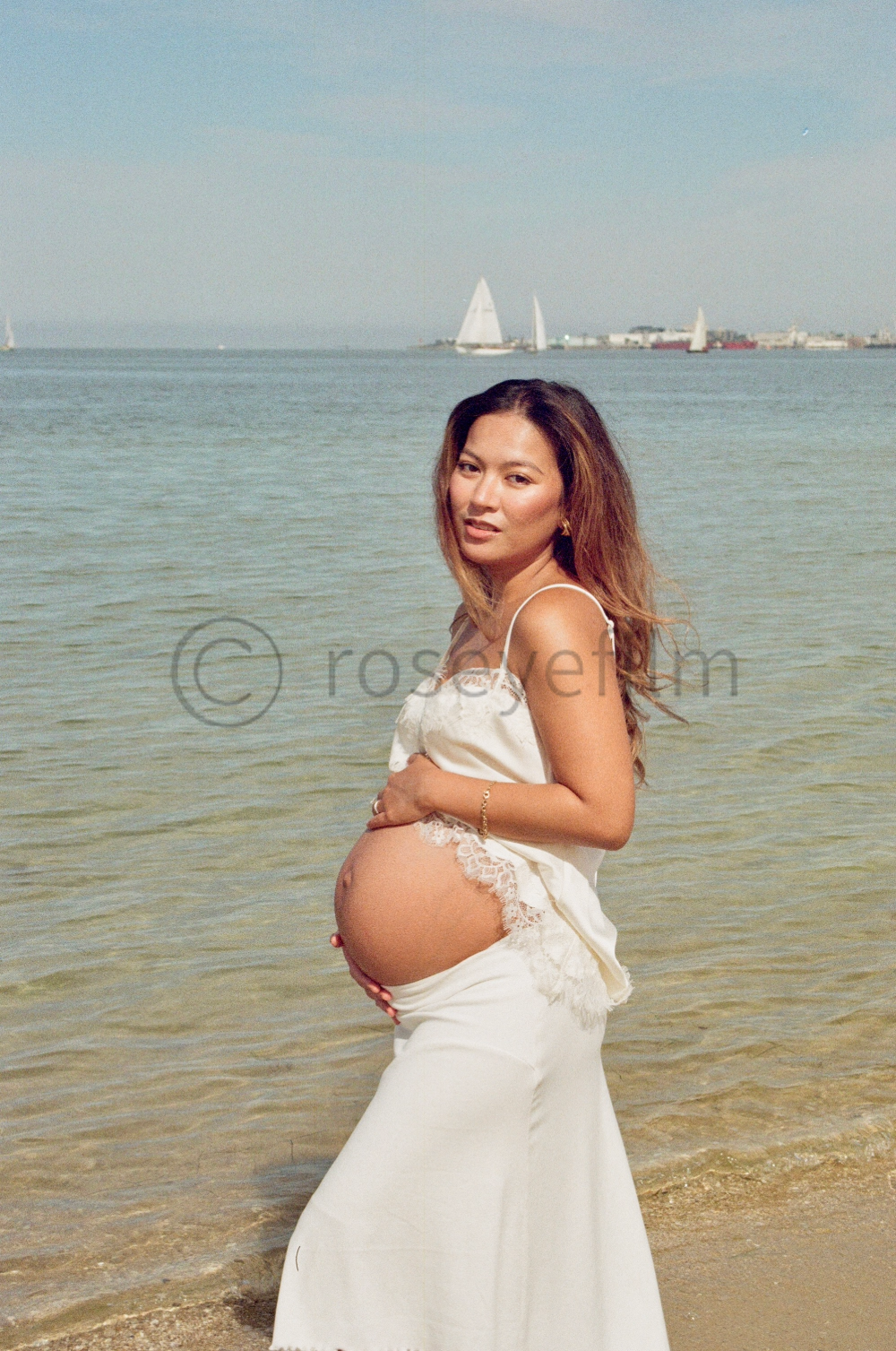 A pregnant woman in a white lace-trimmed outfit standing in the shallow water at the beach with sailboats in the background.