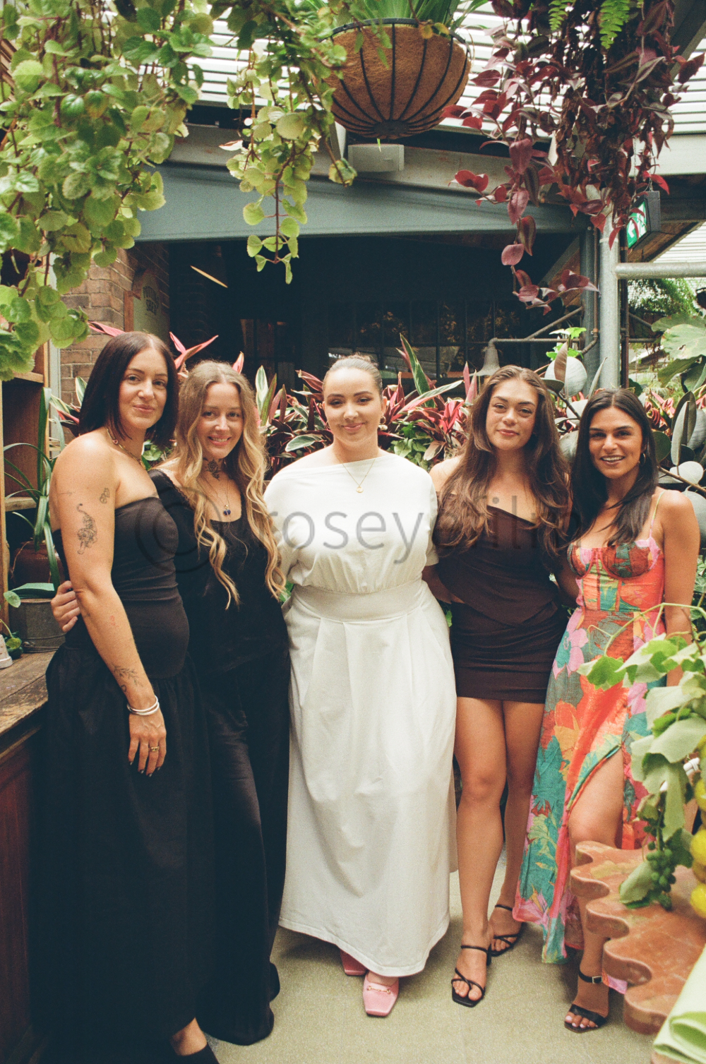 Group of five women standing together in a lush, indoor garden with hanging plants and colorful foliage.