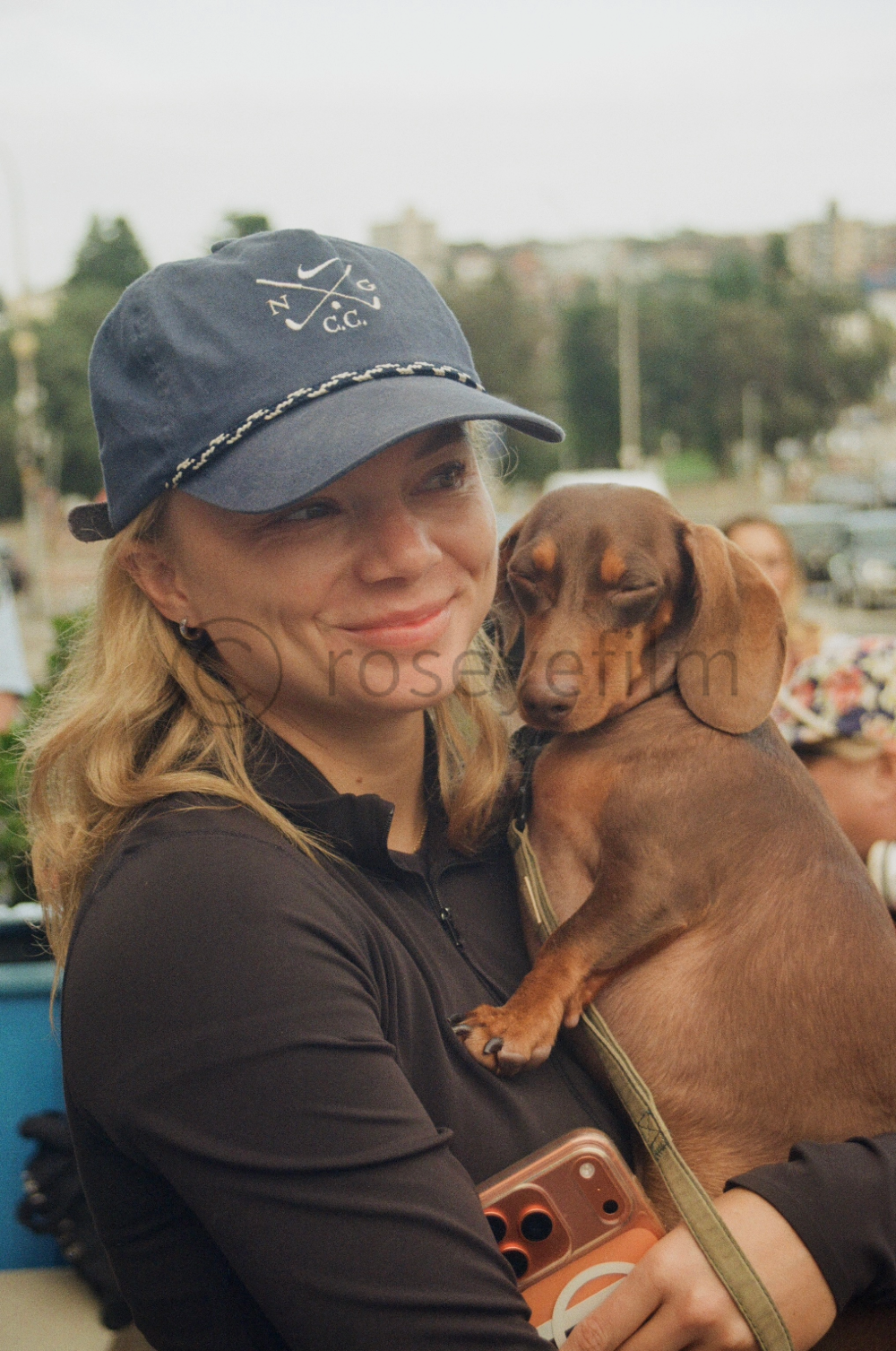 A woman wearing a blue cap and black jacket holding a sleeping brown dachshund dog outdoors.