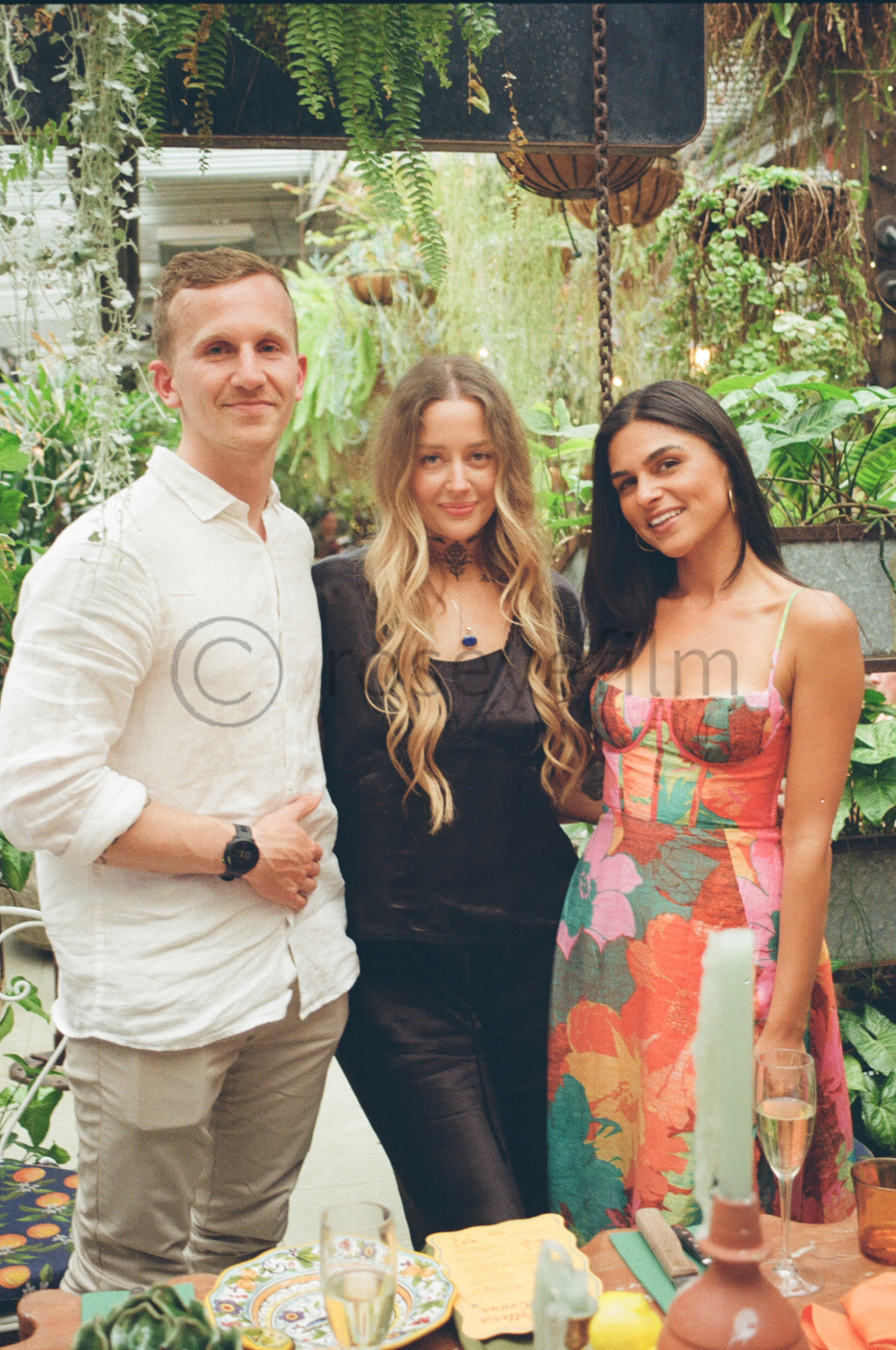 Three people smiling and posing together in a lush, green indoor garden or greenhouse. Two women and one man are standing close to each other, with plants and decorative elements in the background.
