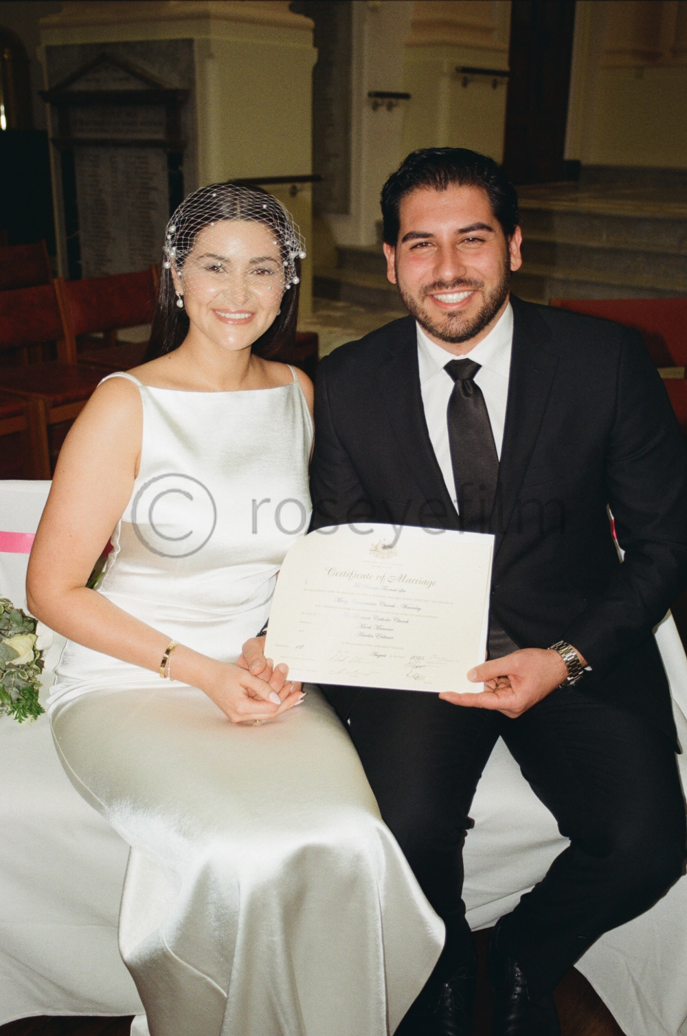 A bride and groom sit together at their wedding, holding a marriage certificate and smiling at the camera inside a church or wedding venue. The bride wears a white satin dress and a birdcage veil, while the groom wears a black suit and tie.