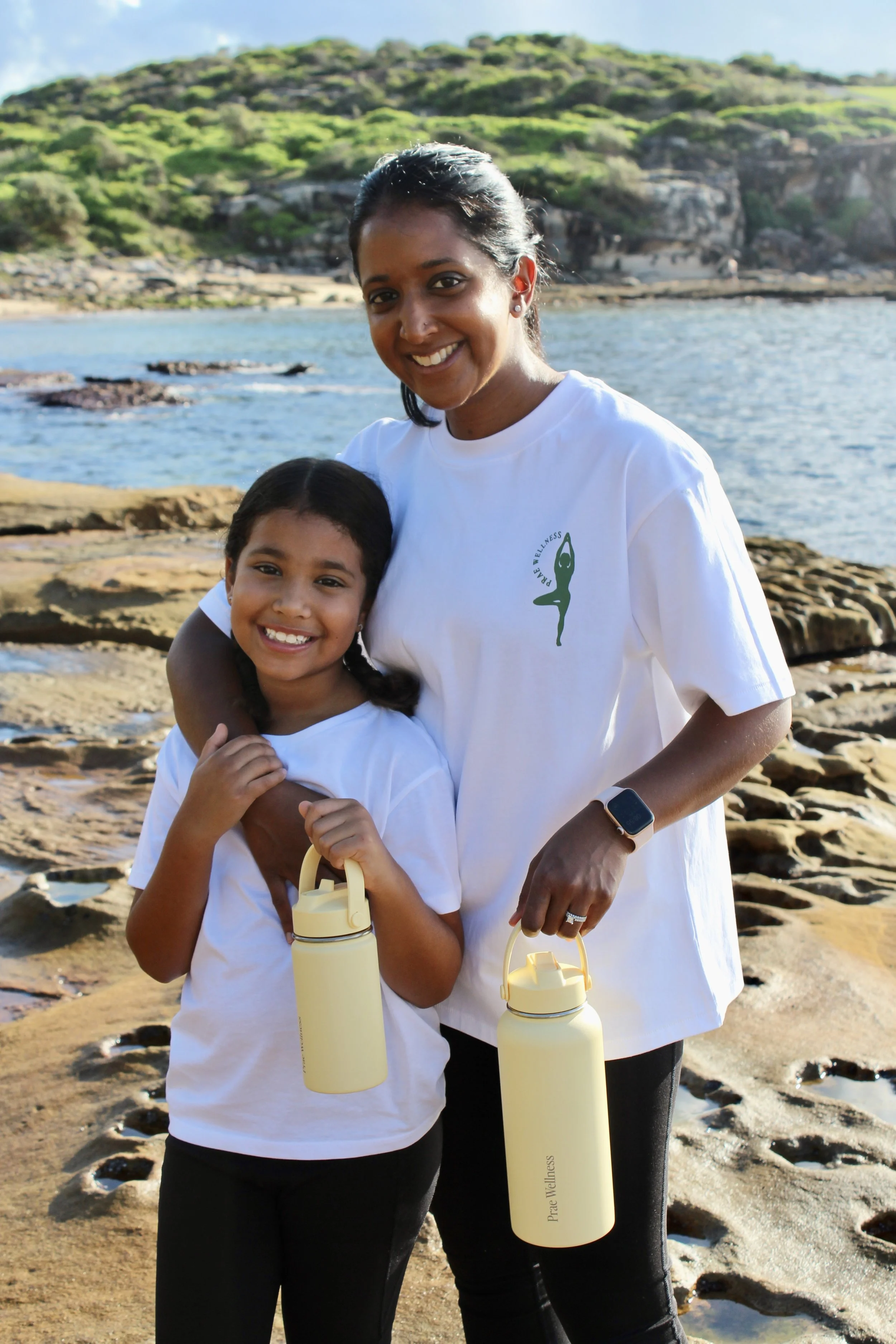 A woman and a girl at the beach, both smiling and holding yellow water bottles, with rocky shoreline and green hills in the background.