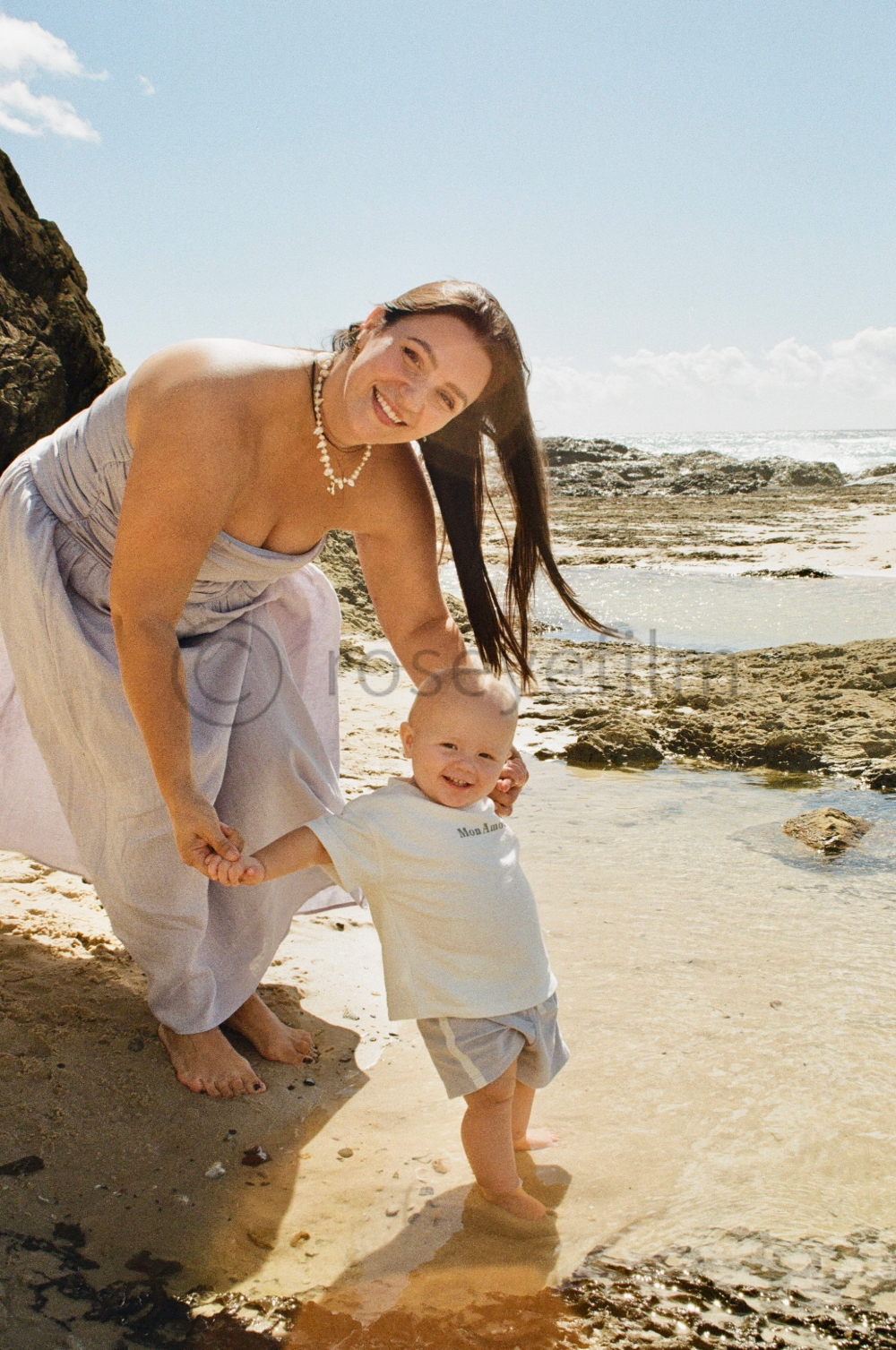 A woman and a toddler playing on a beach near rocks and water, smiling and holding hands.