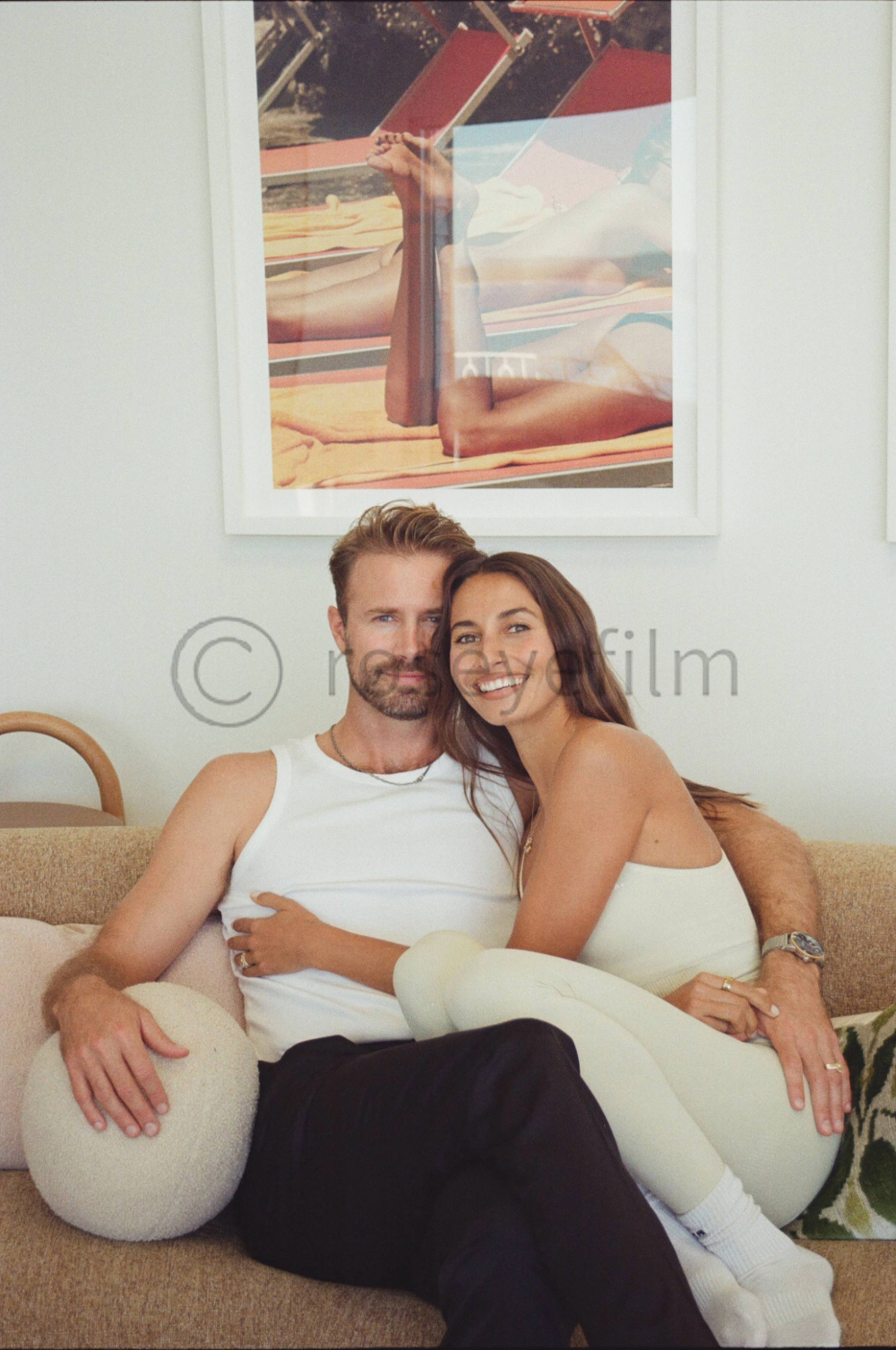 A couple sitting on a beige sofa, embracing each other and smiling at the camera, with a framed picture of a woman sunbathing behind them.